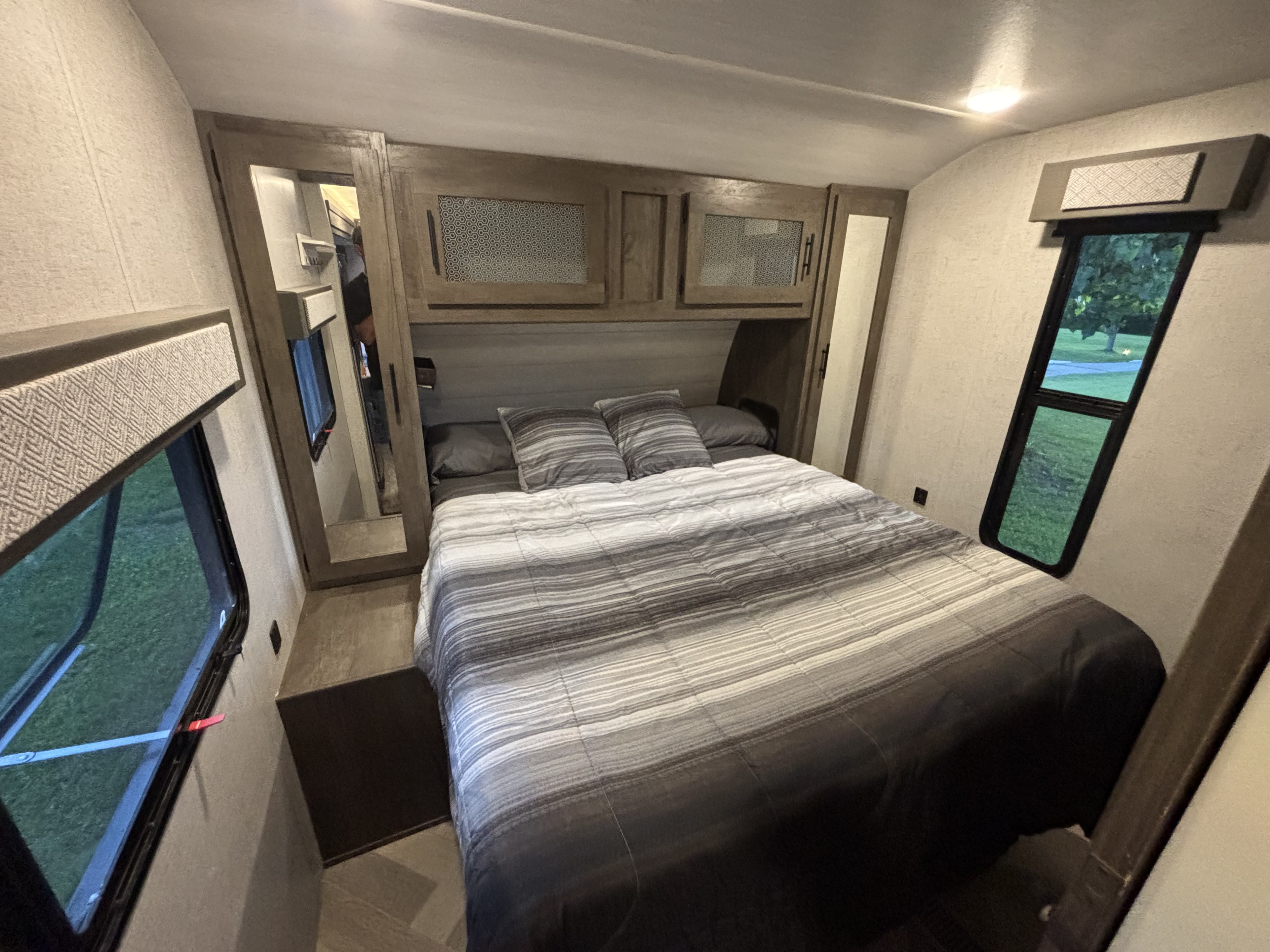 Cozy RV rental bedroom featuring a striped bed, overhead storage cabinets, and two windows offering a glimpse of outdoor greenery.. Forest River Salem 2021