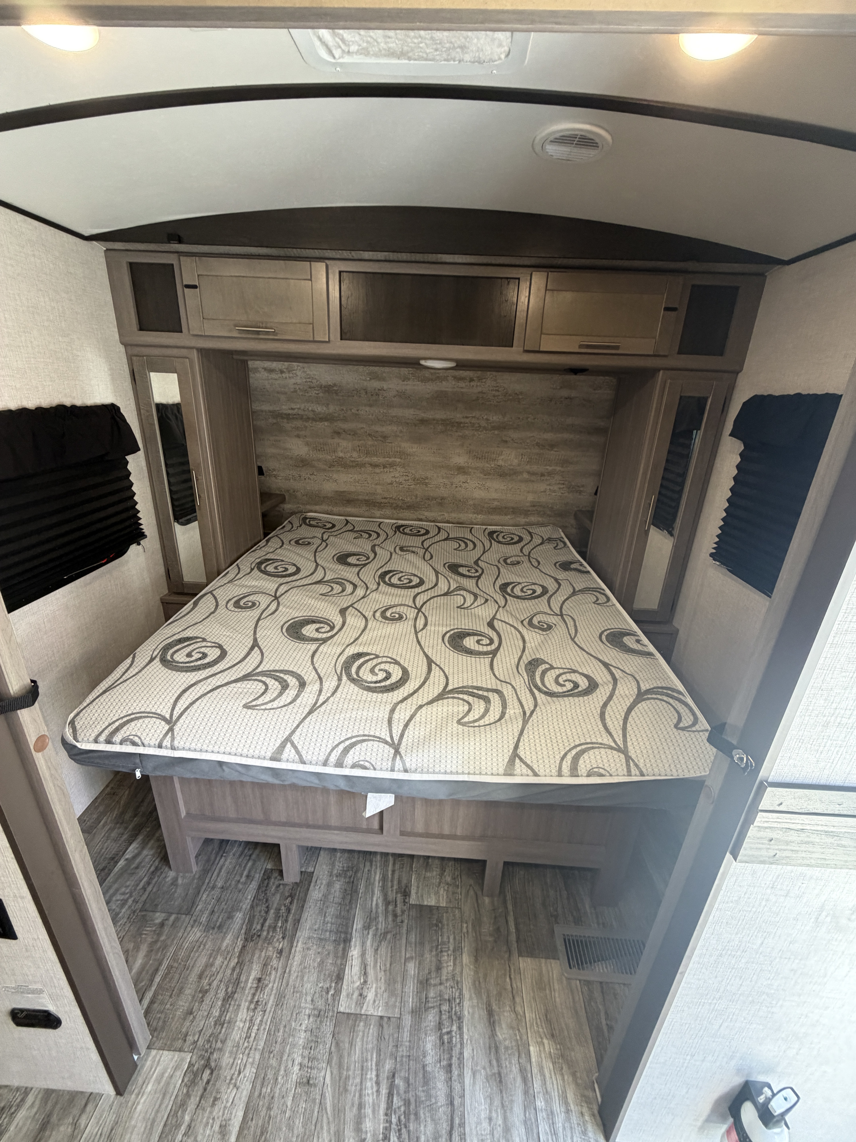 RV rental bedroom with a patterned bed, overhead storage, mirrored closets, black window blinds, and wood-look flooring.. Crossroads RV Sunset Trail Super Lite 2021
