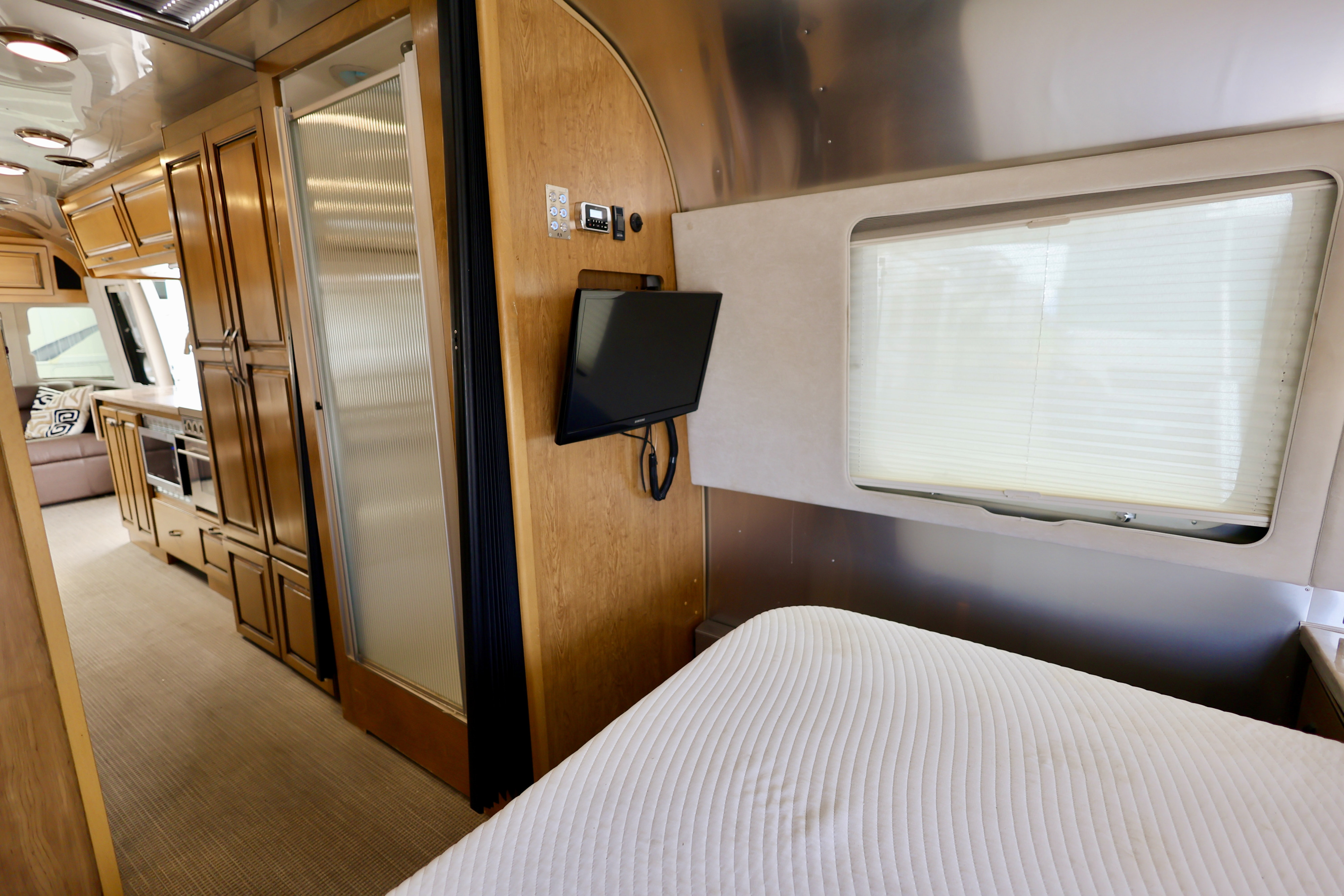 Modern rv rental interior: bedroom with quilted bed, TV, window, leading to sleek wooden cabinetry, shower, and galley.. Airstream Classic 2019