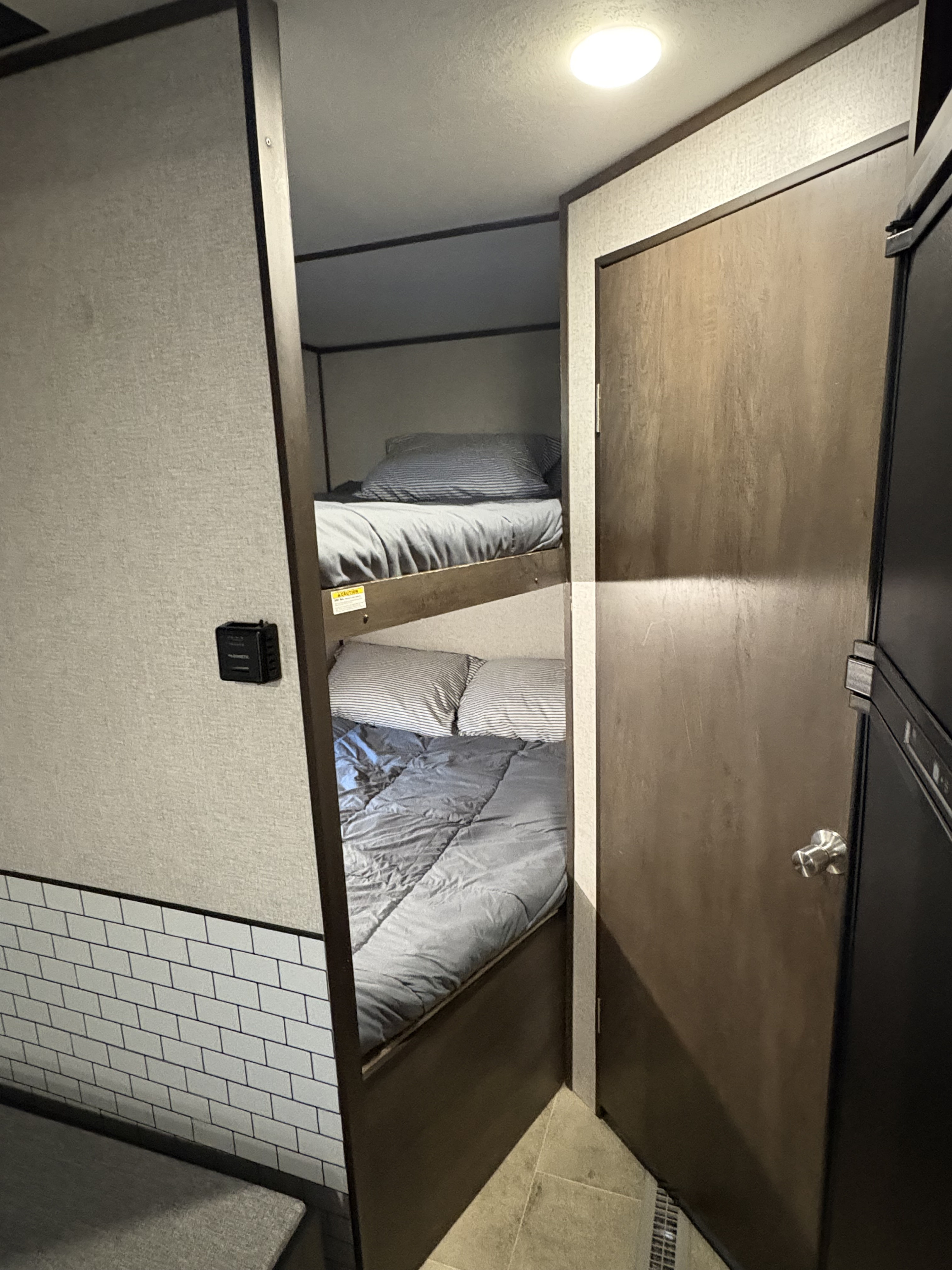 RV rental interior: bunk beds with gray bedding, a light fixture on the ceiling, a wooden door, and a wall-mounted control panel.. Dutchmen Coleman Lantern LT 2020