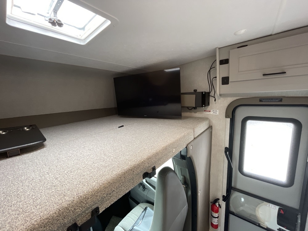 Inside an RV rental: view of the cab-over bunk with a TV and skylight, leading to the entrance door.. Coachmen Freelander 22xg 2024