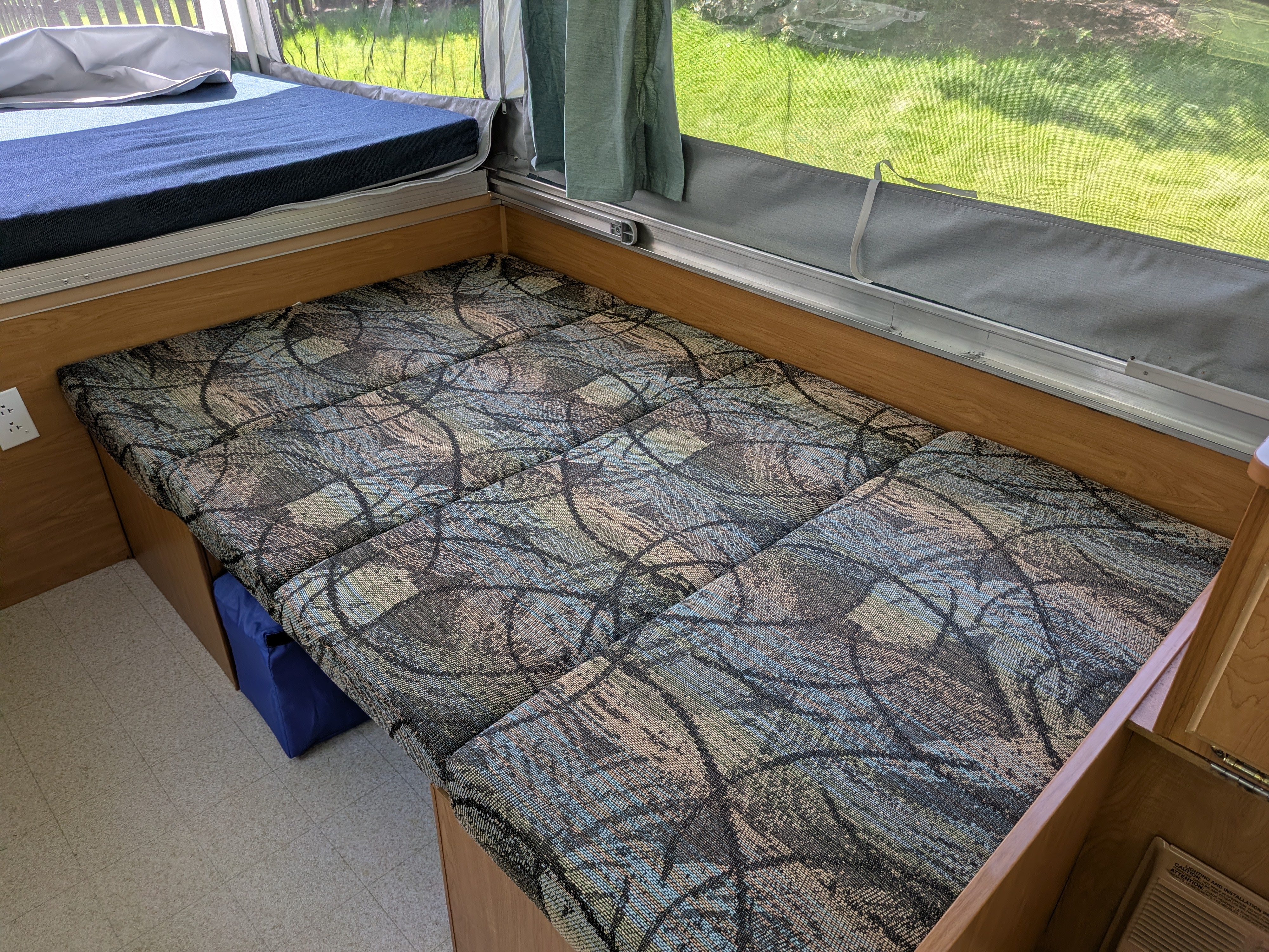 Interior view of an RV rental showing a blue mattress on a bed, colorful patterned dinette cushions, and a screened window.. Coleman Destiny Sedona 2002