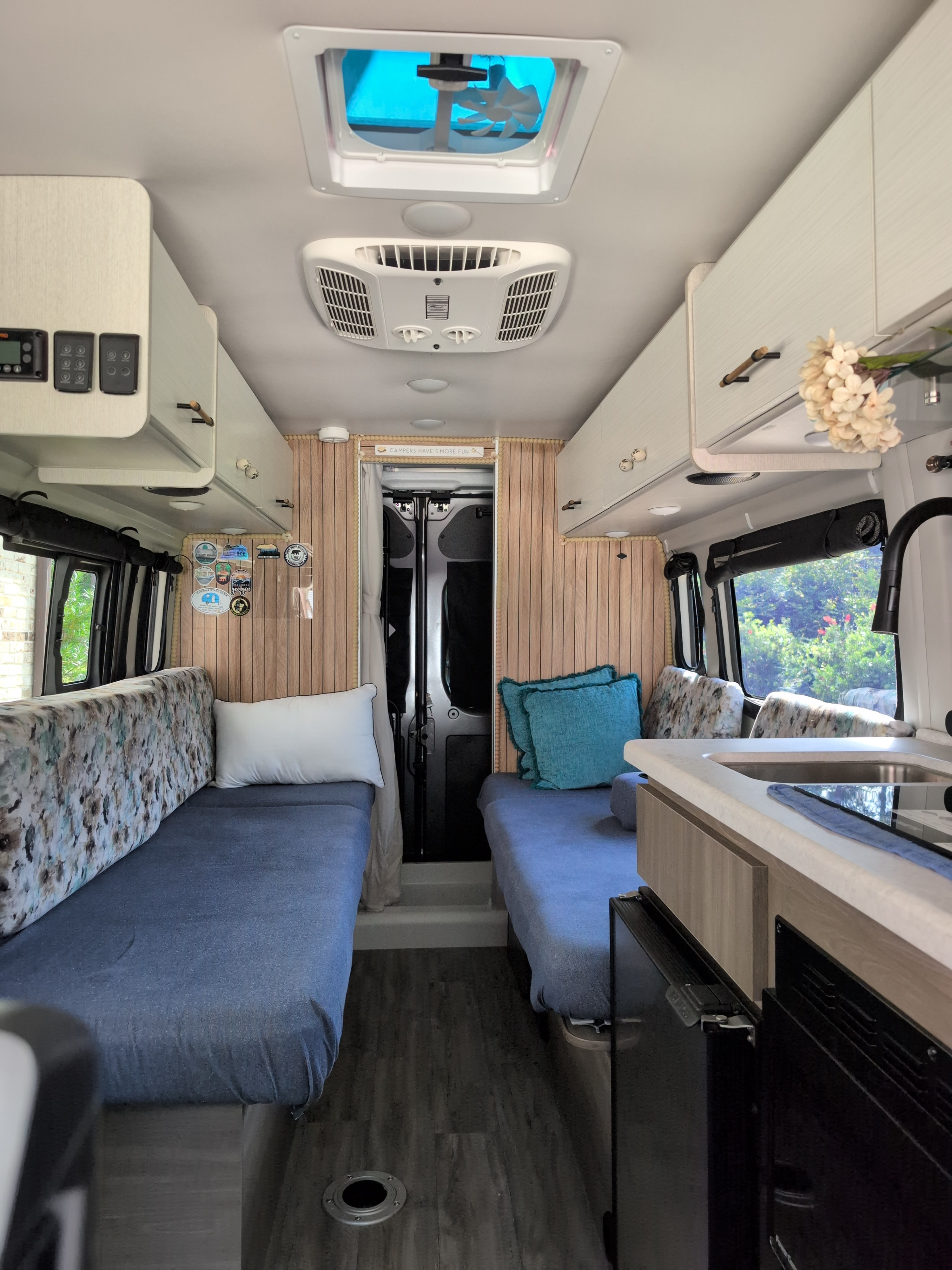 Bright rv rental interior: two blue-covered twin beds, kitchen counter, overhead cabinets, wooden walls, and ceiling fan/AC. Cozy camper setup.. Thor Tellaro 2024