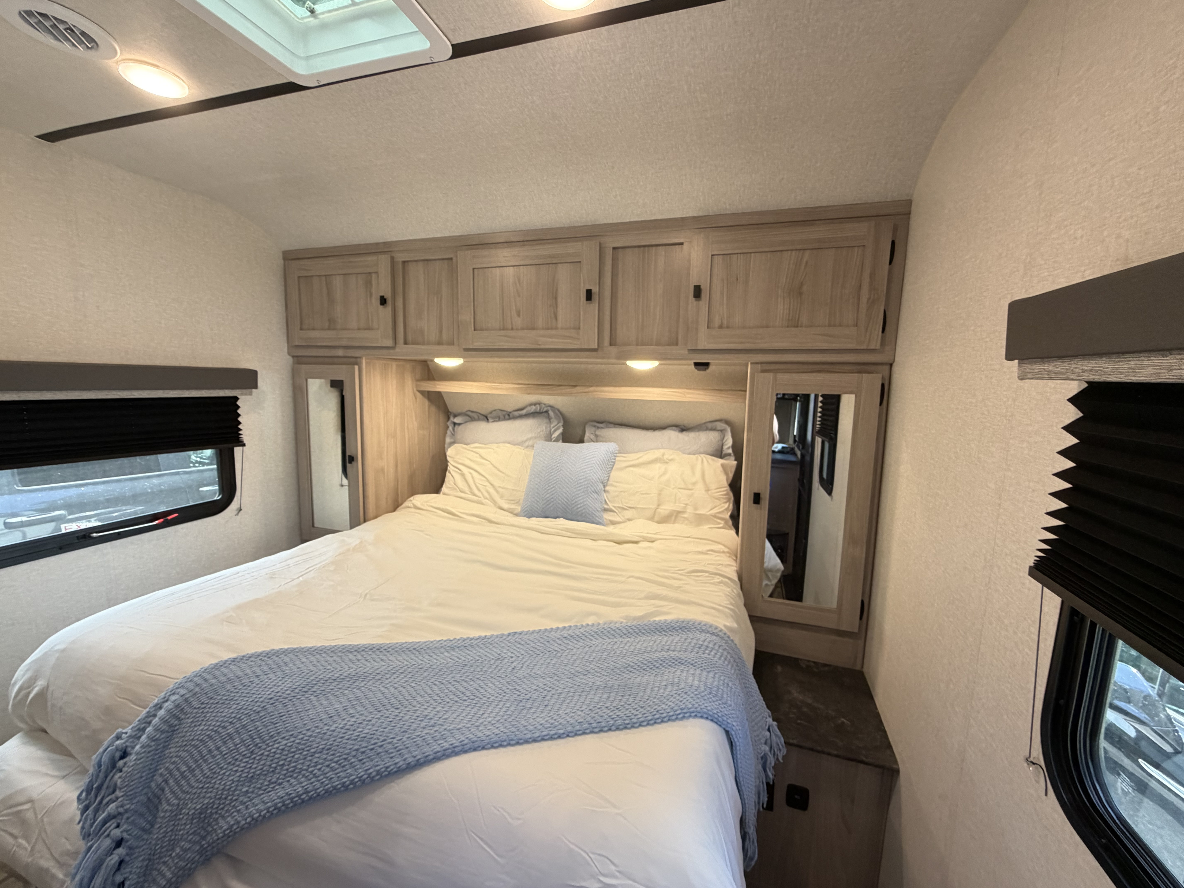 Cozy RV rental bedroom with a comfortable queen bed, light wood cabinets, mirrors, and windows with blinds.. Coachmen 2963BH 2023