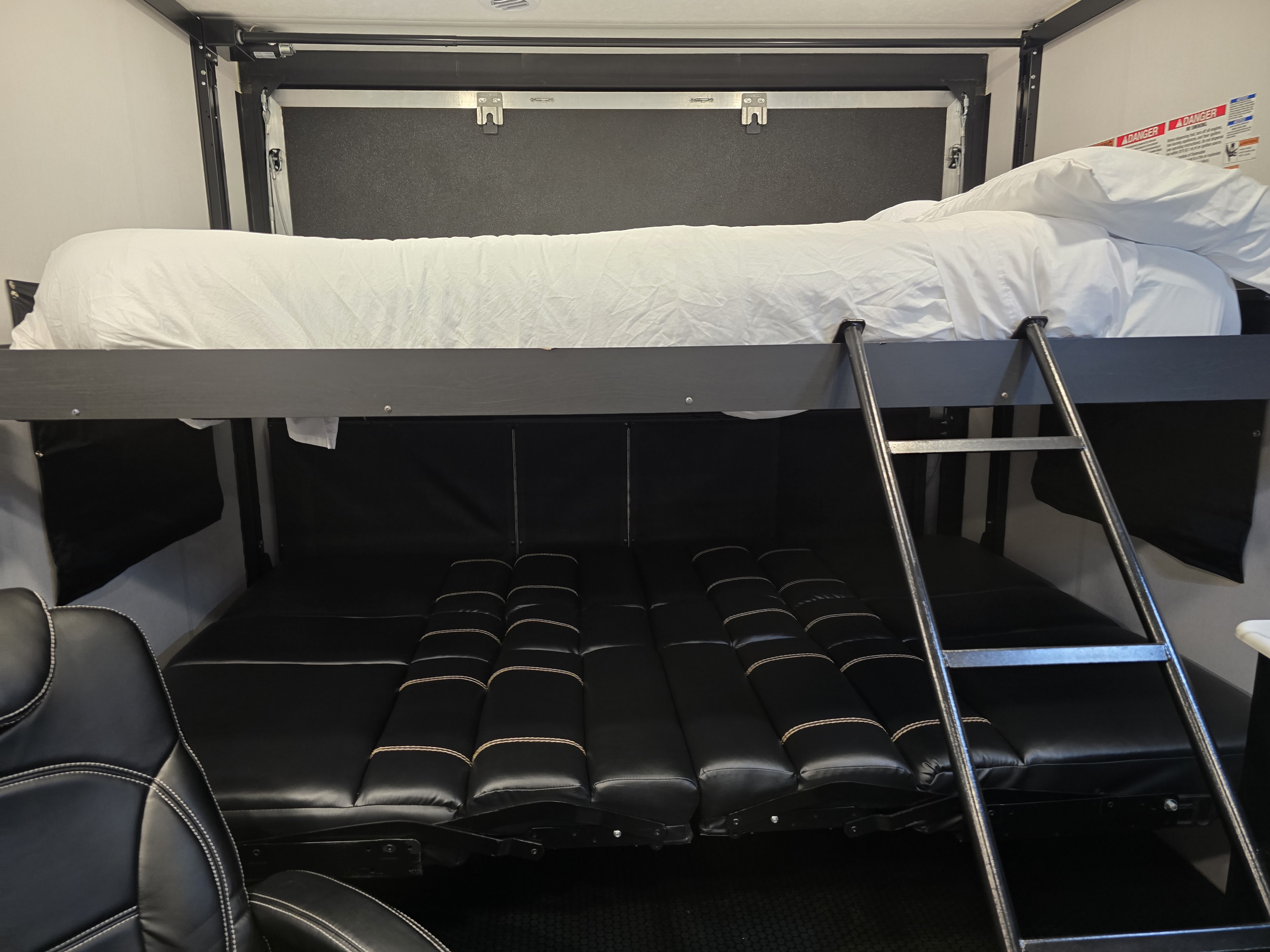 RV rental interior featuring a bunk bed with white sheets, a ladder, and a lower convertible black padded seating/bed area.. Coachmen Adrenaline 2022
