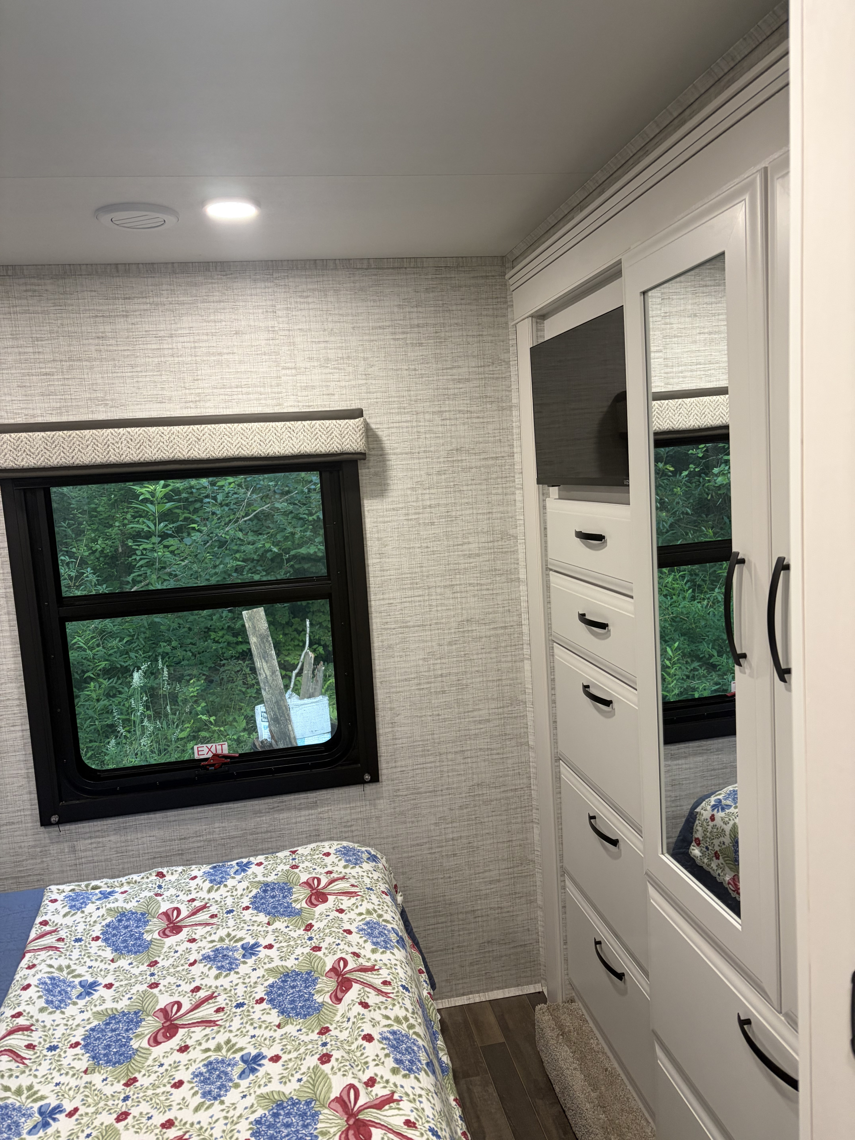 Cozy RV rental bedroom with a floral bedspread, window overlooking green trees, white wardrobe, built-in TV, and modern decor.. Entegra Coach Odyssey 2025