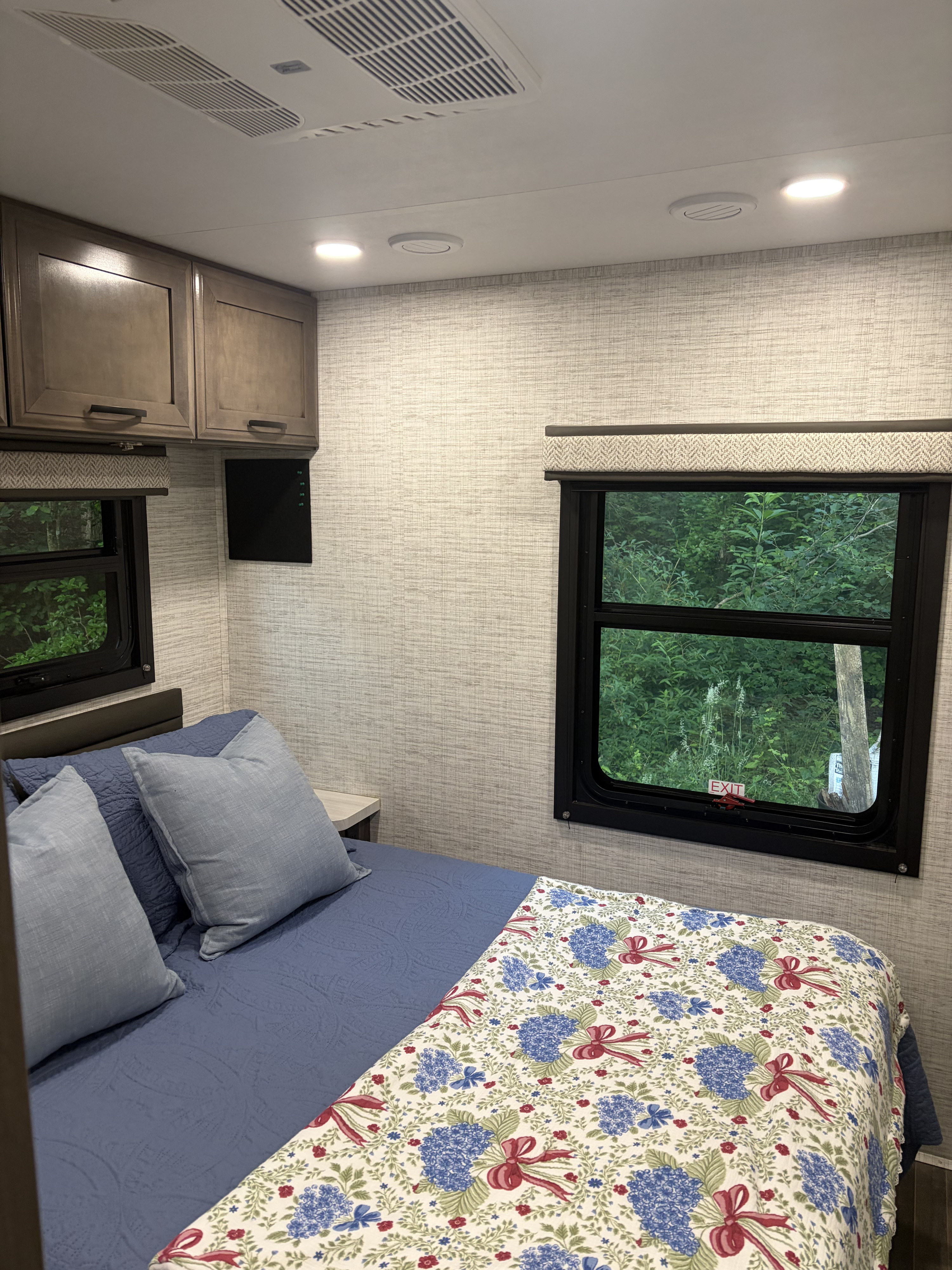 Cozy RV rental bedroom with blue bed, patterned quilt, wall cabinets, and windows offering a view of lush greenery.. Entegra Coach Odyssey 2025