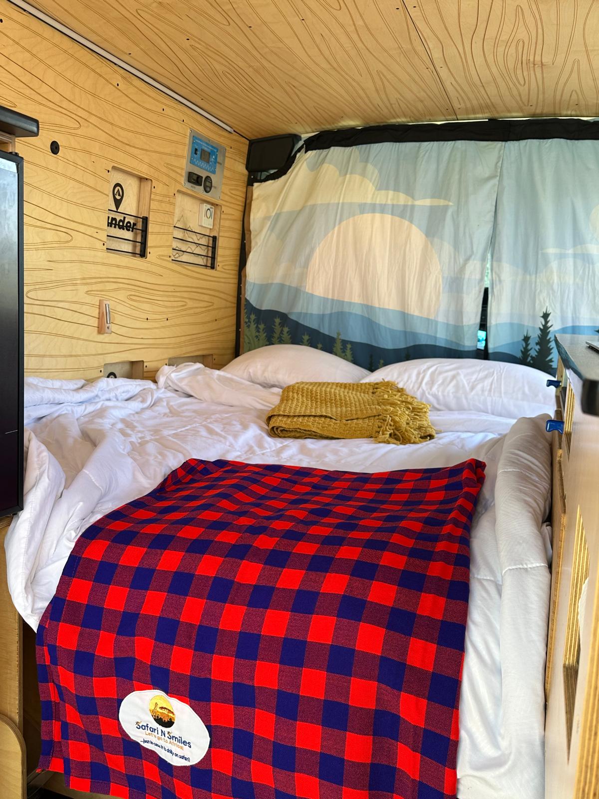 Inside a cozy **rv rental**: a comfortable bed with red/blue plaid blanket, white bedding, and scenic nature-themed curtains.. RAM Promaster 2023