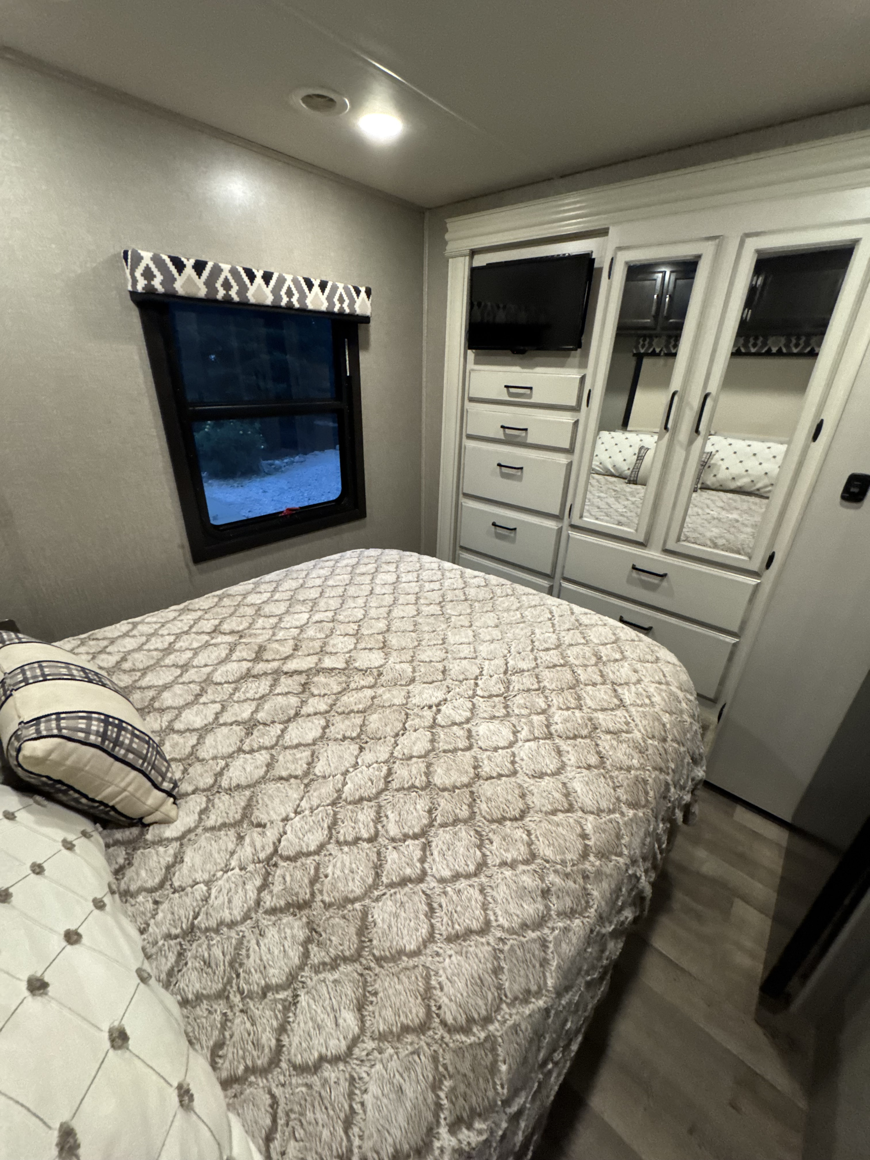 Cozy RV rental bedroom featuring a comfy bed with textured comforter, window, and built-in wardrobe with TV.. Jayco Greyhawk 2021