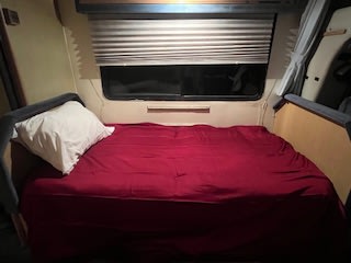 Inside an RV rental, a bed with red sheets and a white pillow awaits, under a window with closed blinds.. Winnebago Warrior 1990