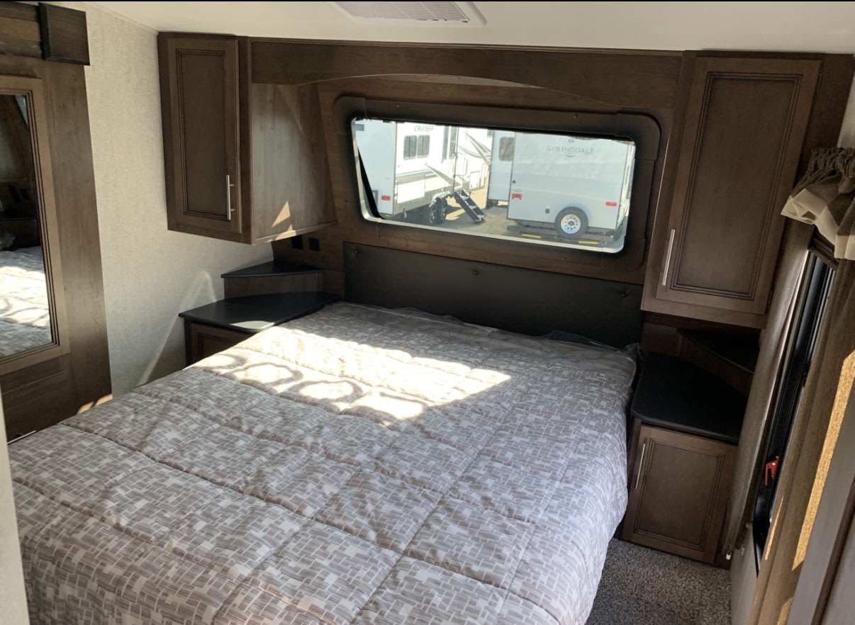 **RV rental** bedroom interior: bed with patterned comforter, dark wood cabinets, and window overlooking other travel trailers.. Keystone RV Cougar 2020