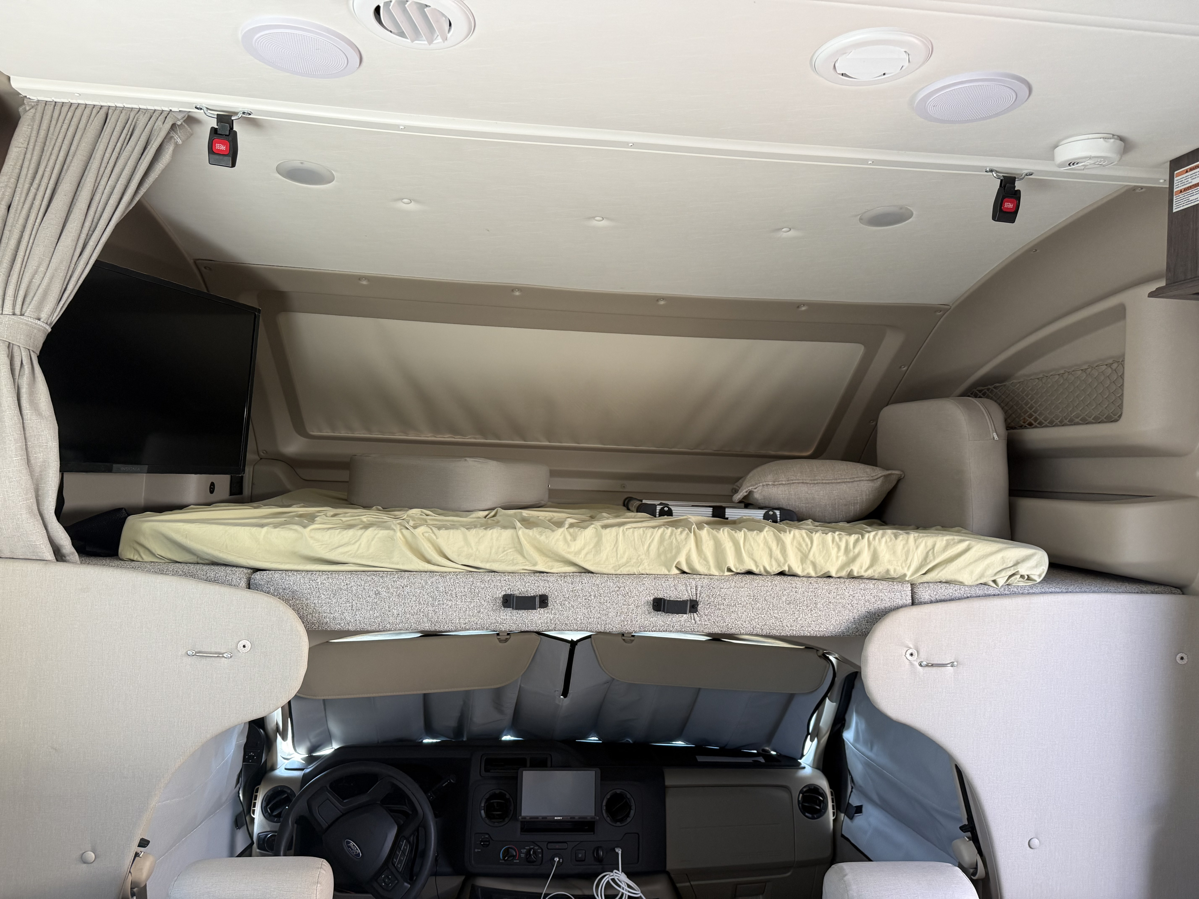 RV rental overhead bunk bed with a TV, pillows, curtains, and a view of the dashboard from above, including a ceiling seatbelt.. Entegra Coach Esteem 2023