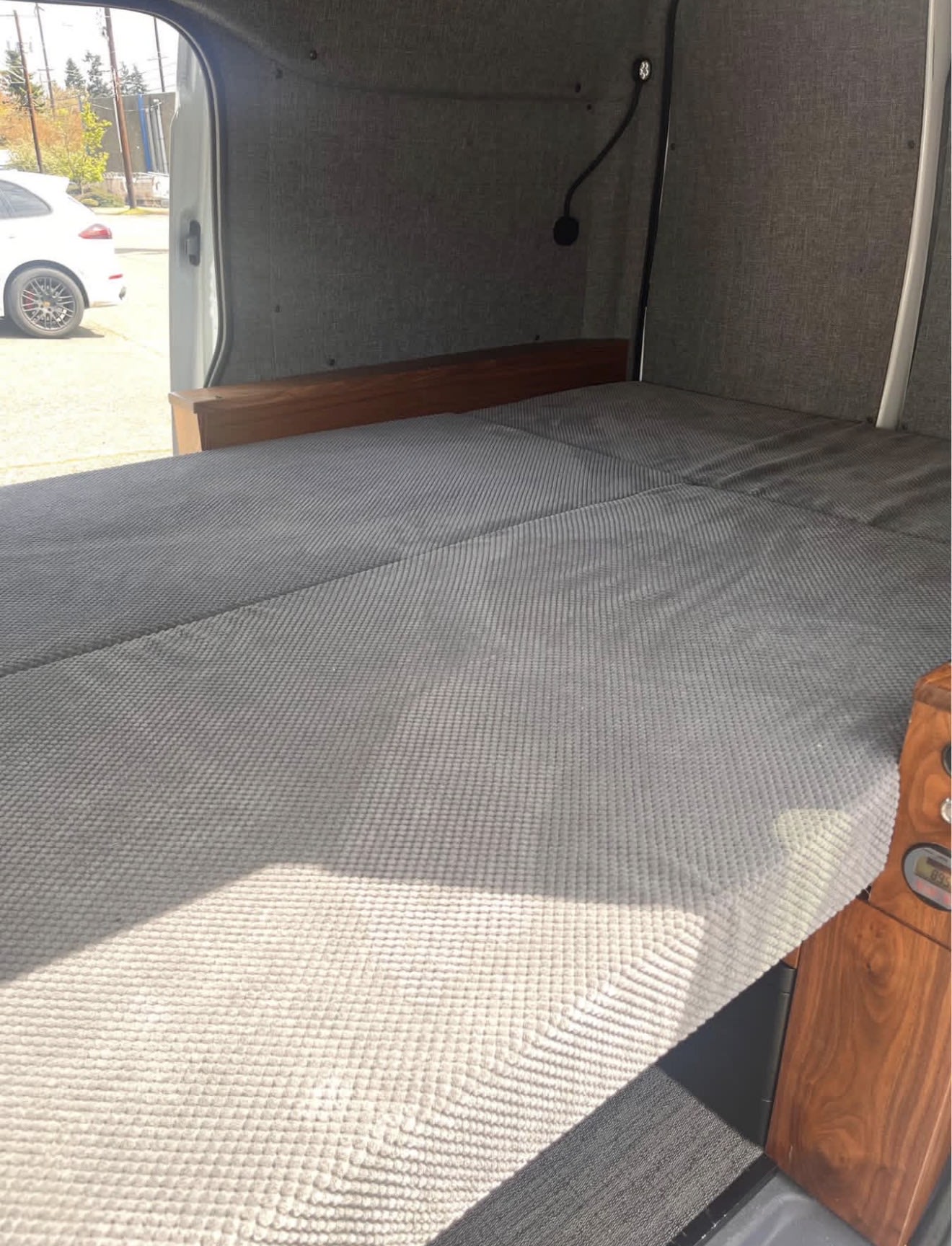RV rental interior: A comfortable grey textured bed and wooden accents inside a camper van, with the door open to the outdoors.. Ford Transit 2013
