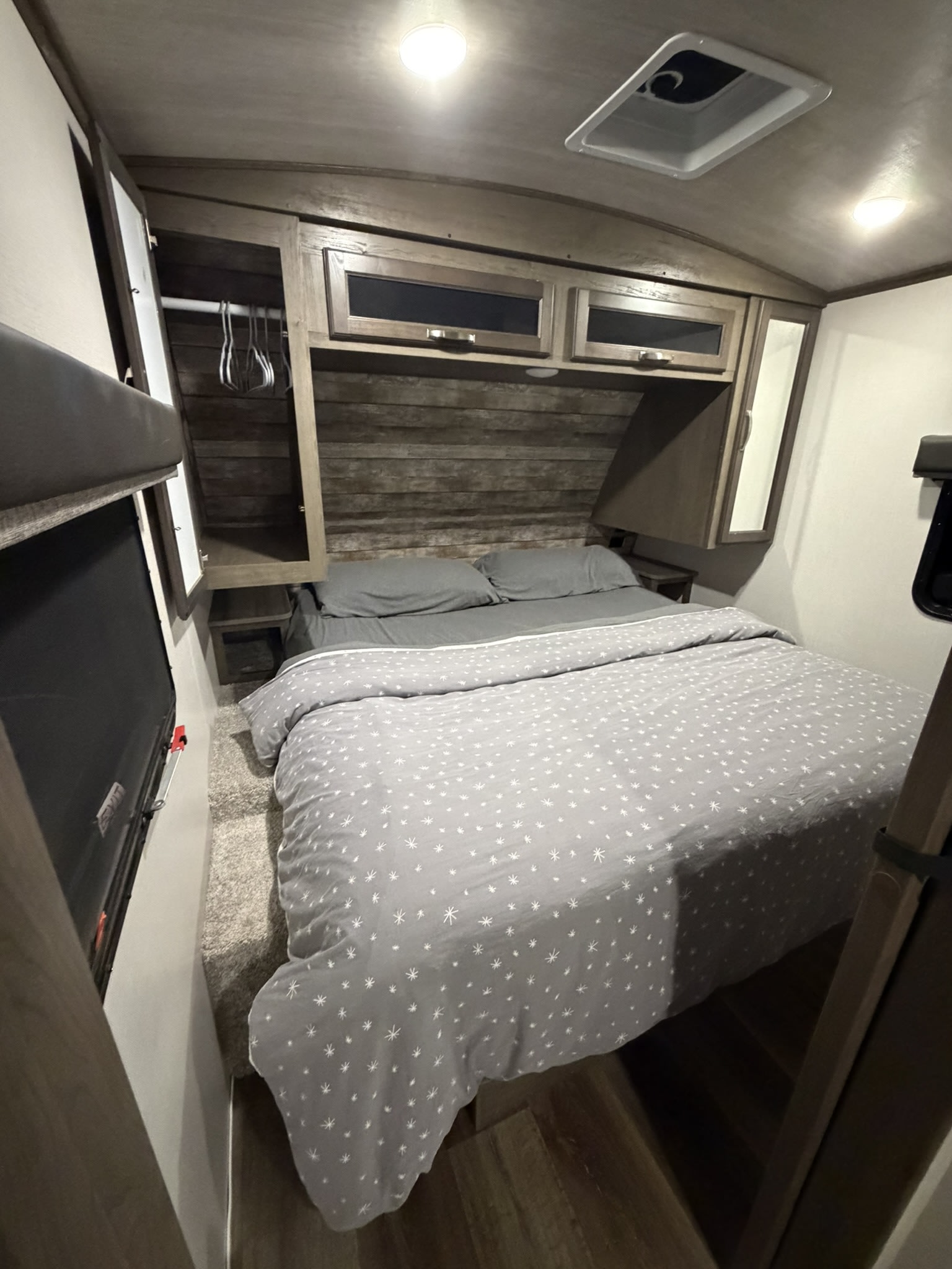 Cozy RV rental bedroom featuring a bed with grey patterned bedding, wood paneling, overhead cabinets, and a ceiling vent.. Keystone RV Cougar 2023