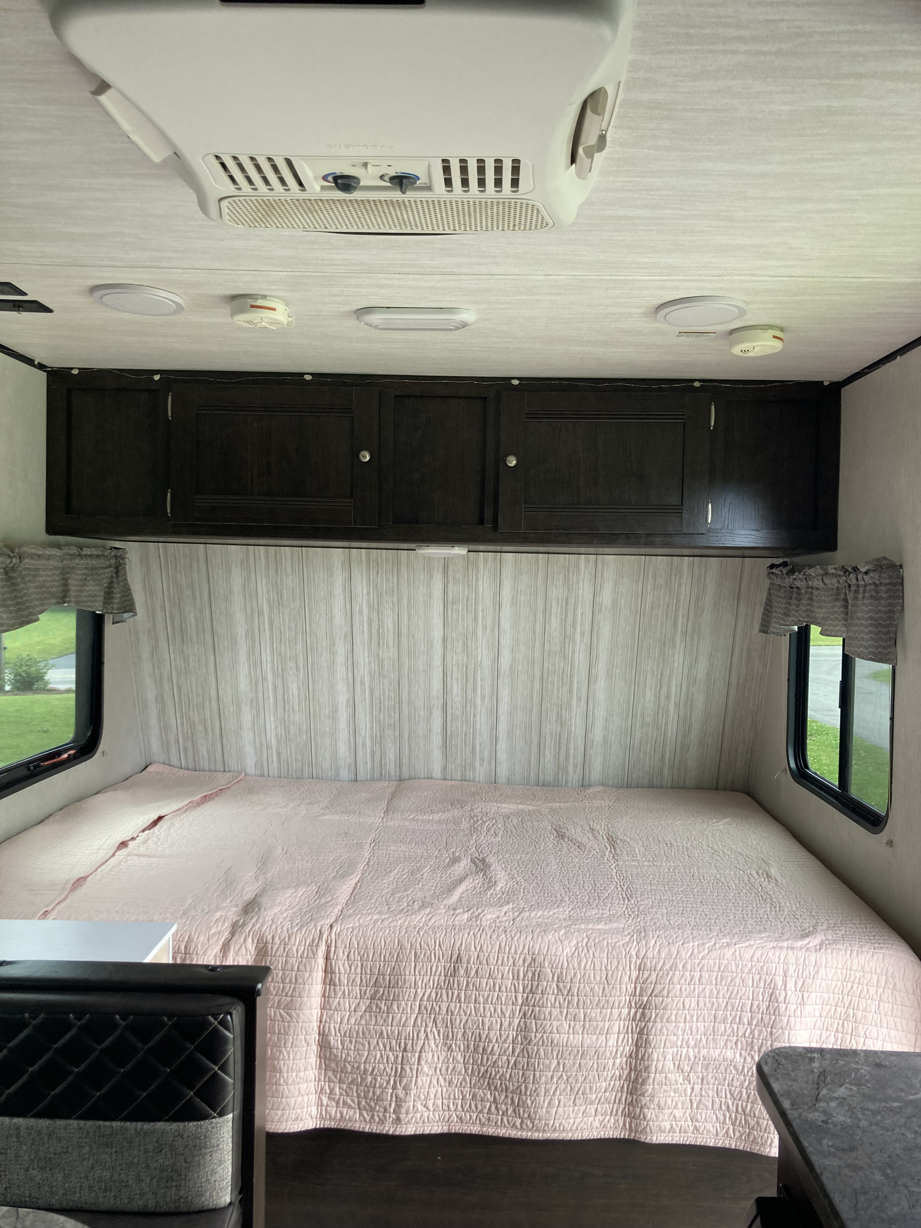 Inside an RV rental: a comfortable bedroom area with a pink quilted bed, dark cabinets, AC, and bright windows.. Heartland RVs Pioneer 2019