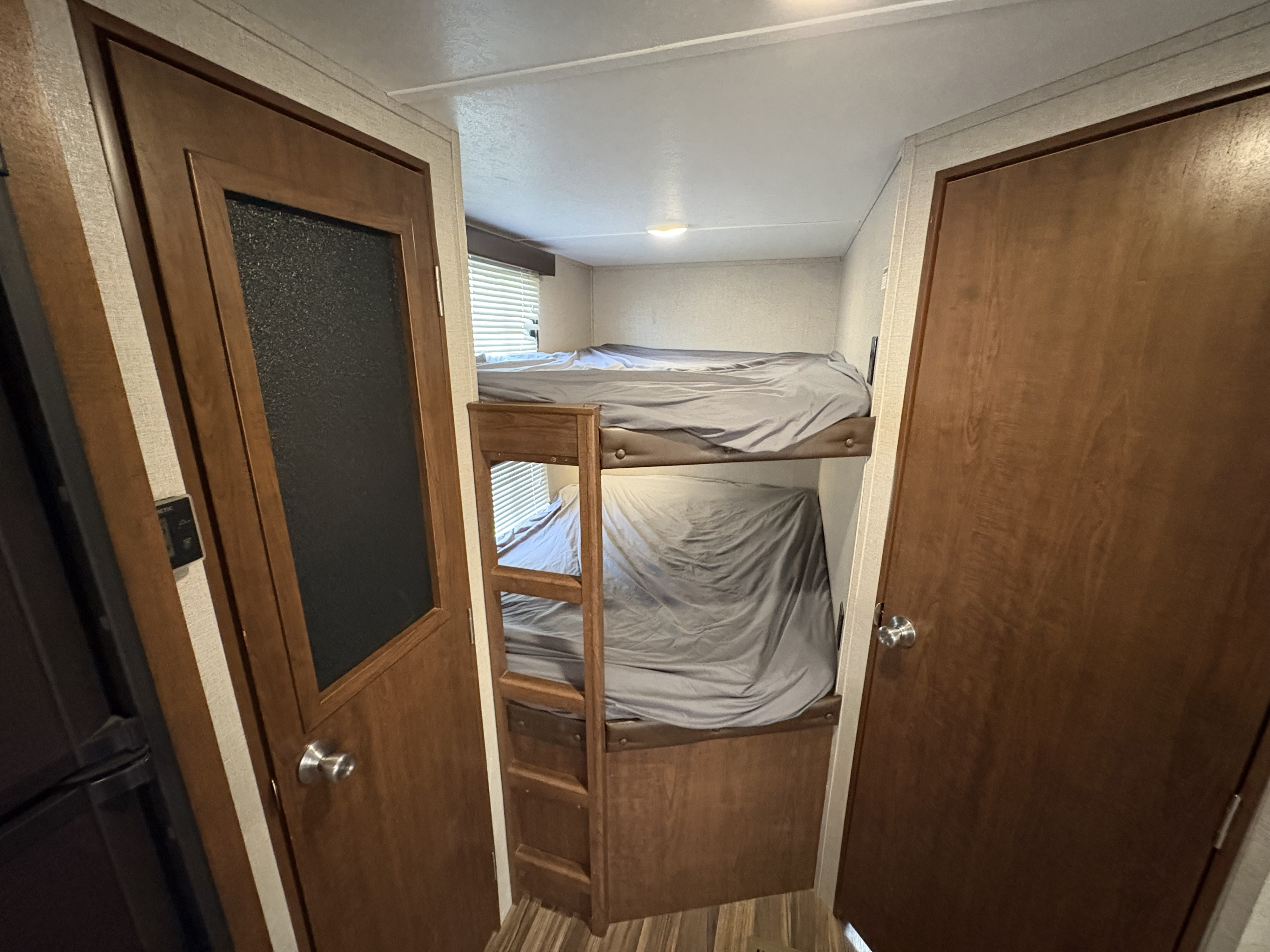 Cozy bunk beds with grey bedding in an RV rental, framed by wooden doors. A window and light add comfort to the compact space.. Keystone RV Hideout 2018