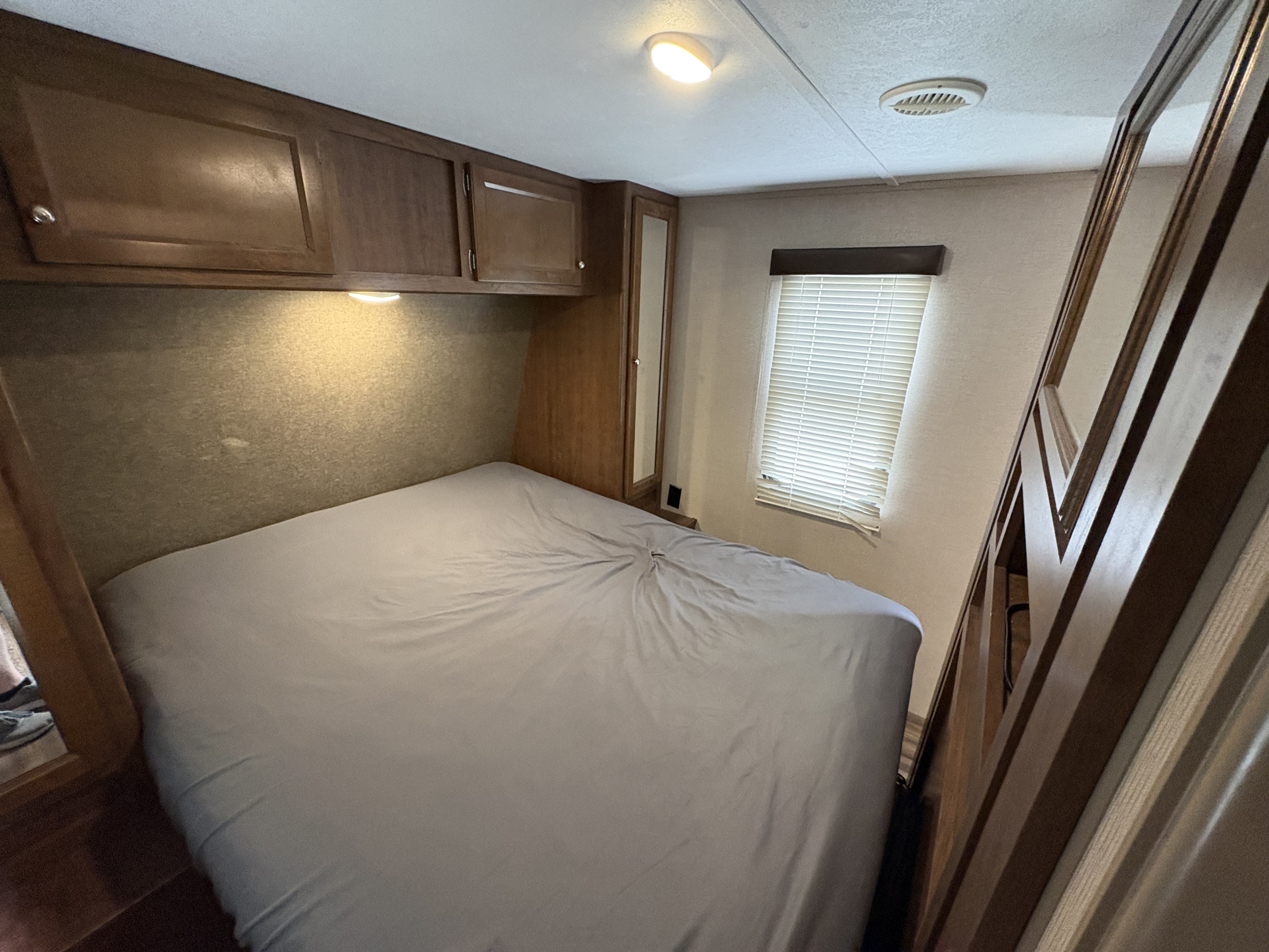 This RV rental bedroom features a cozy bed with overhead wood cabinets, a window with blinds, and soft lighting.. Keystone RV Hideout 2018