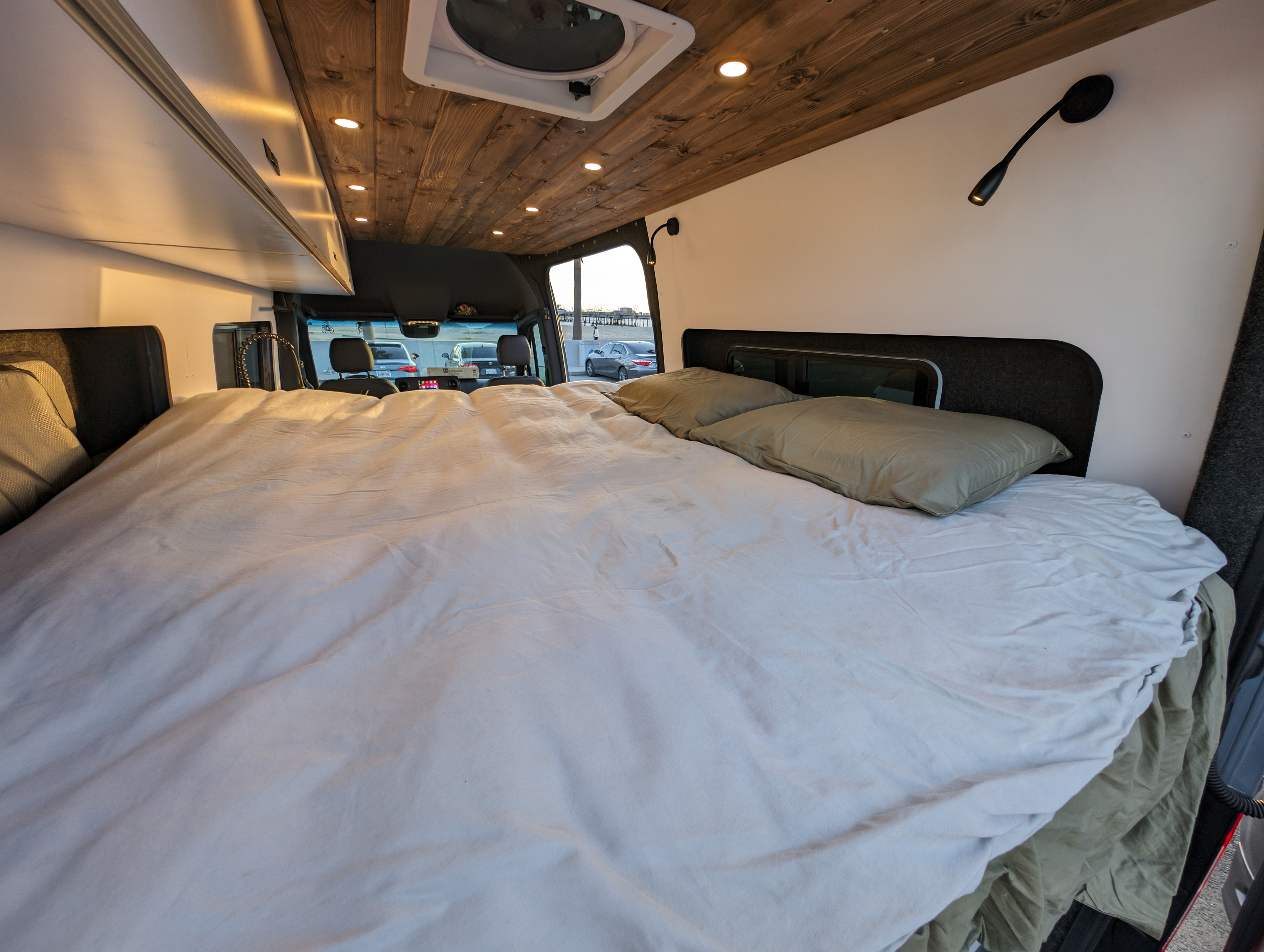 RV rental interior: comfy bed with light bedding, wooden ceiling with lights, and a scenic view of cars and a distant pier.. Mercedes-Benz Sprinter 2023