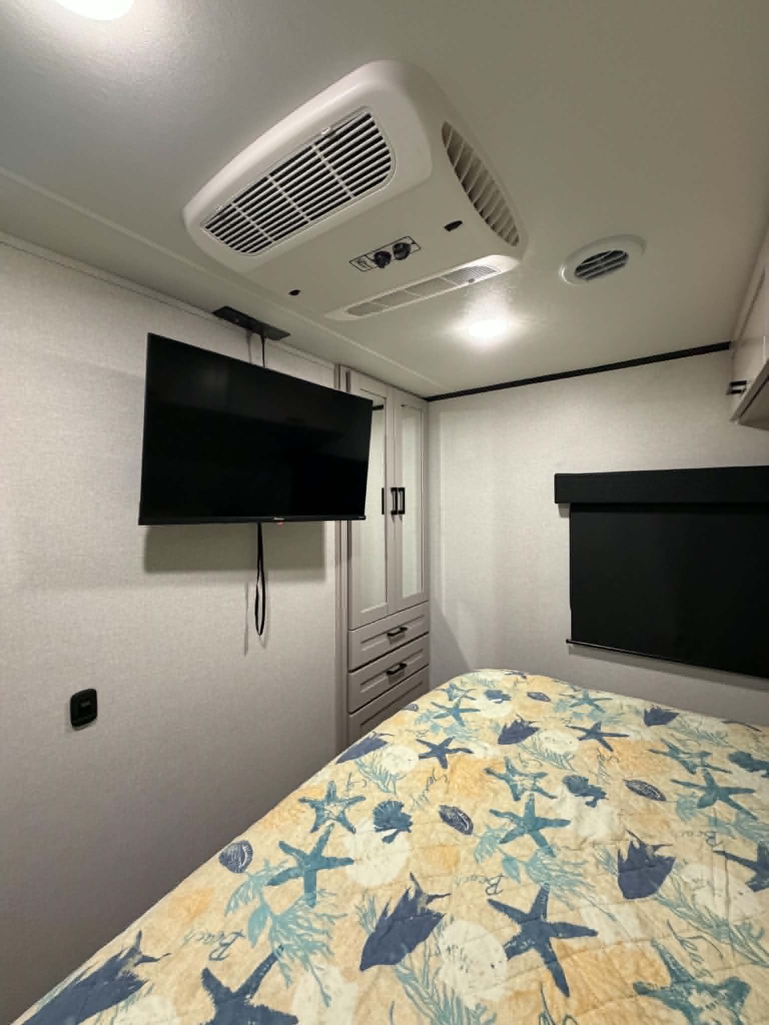 RV rental bedroom with a nautical-themed bed, wall-mounted TV, ceiling AC, and built-in storage cabinets.. Jayco Jay Feather 2025