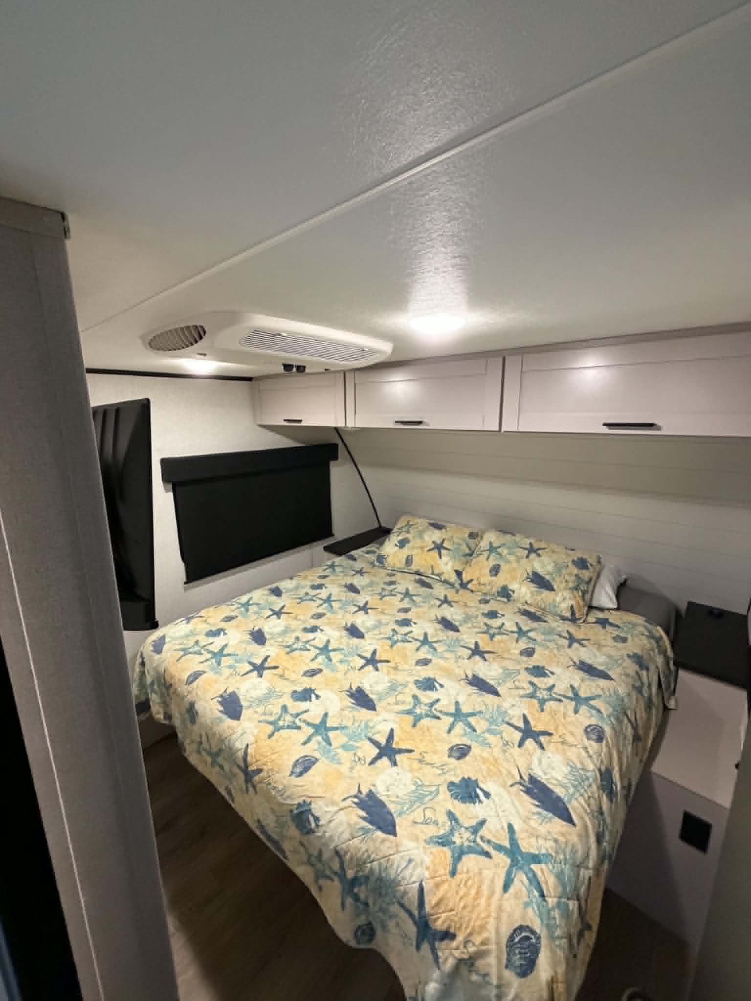 Relax in this RV rental bedroom featuring a charming starfish bedspread, overhead storage, and dark window blinds.. Jayco Jay Feather 2025