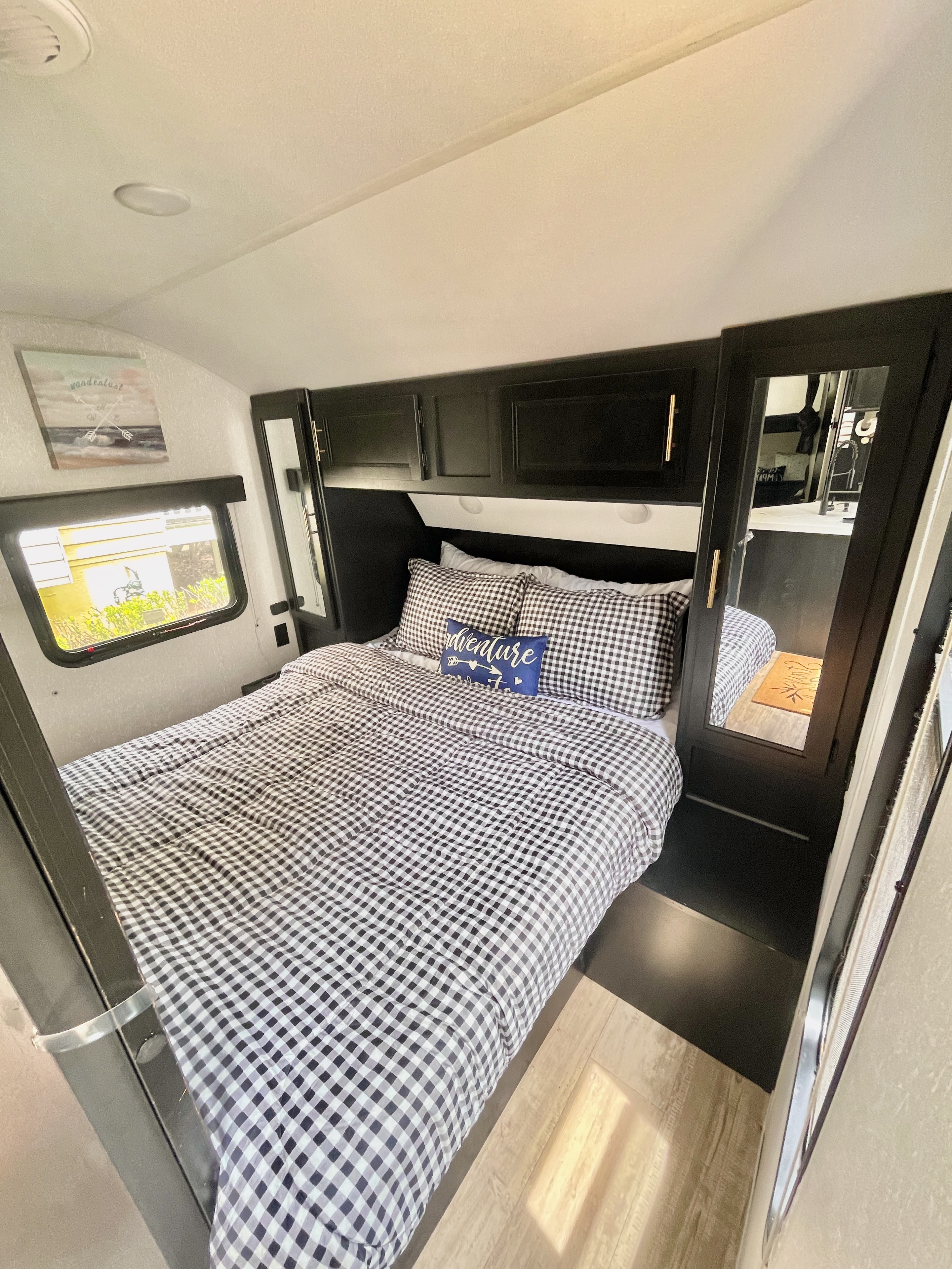 Cozy RV rental bedroom featuring black and white gingham bedding, an "adventure" pillow, dark overhead storage, and a bright window.. Forest River Cherokee Grey Wolf 2021