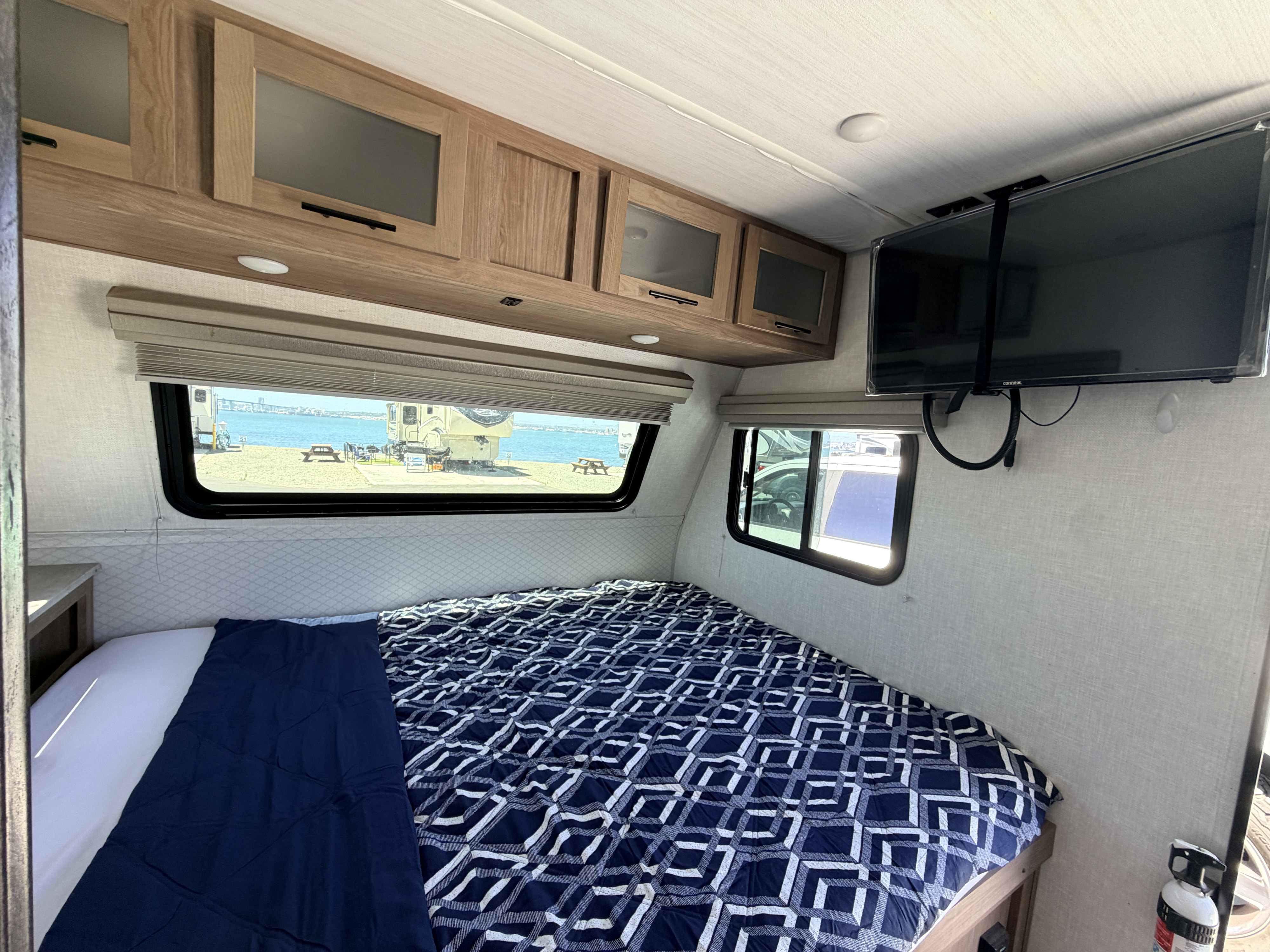 Scenic rv rental interior: bedroom with patterned bedding, overhead cabinets, mounted TV, and stunning water views.. No Boundaries 19 2021