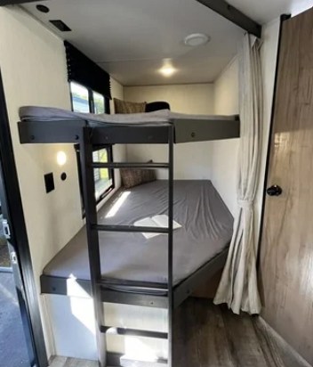 This RV rental offers a compact bunk bed sleeping area. Features a ladder, gray mattresses, window, and cozy lighting.. Forest River Campsite Reserve 23LM 2025