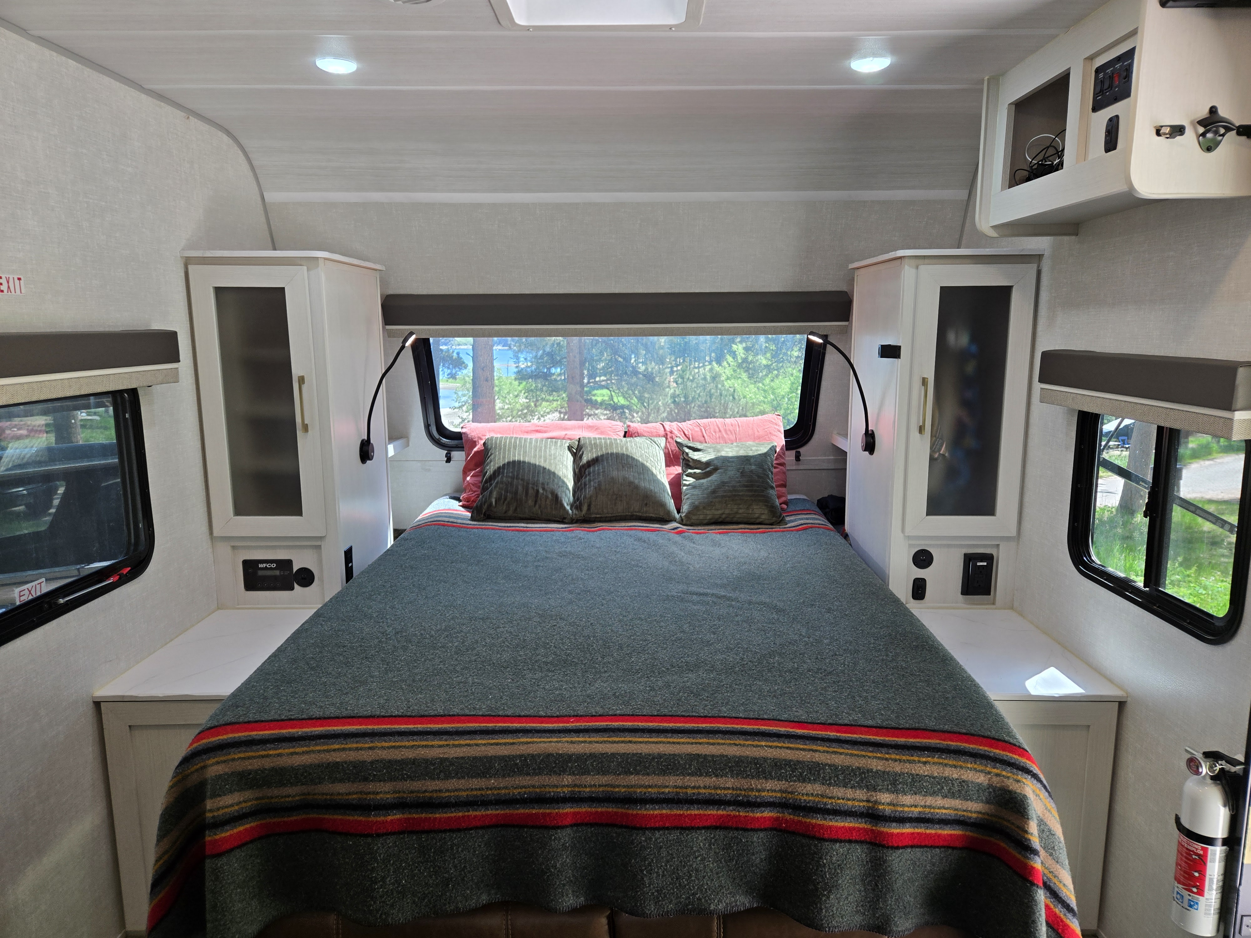 A bright and cozy RV rental bedroom features a bed with a striped blanket, overhead lighting, and scenic views from windows.. No Boundaries 20 2025