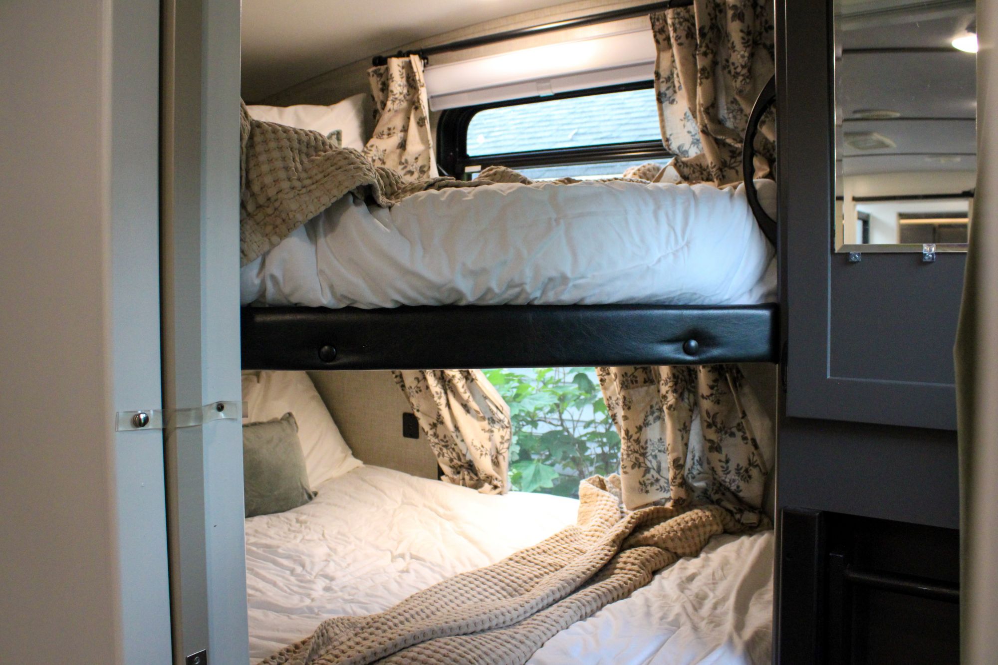 Cozy bunk beds in an RV rental, featuring white bedding, a textured blanket, and floral curtains by the windows. Green foliage visible.. Keystone RV Outback Ultra-Lite 2021