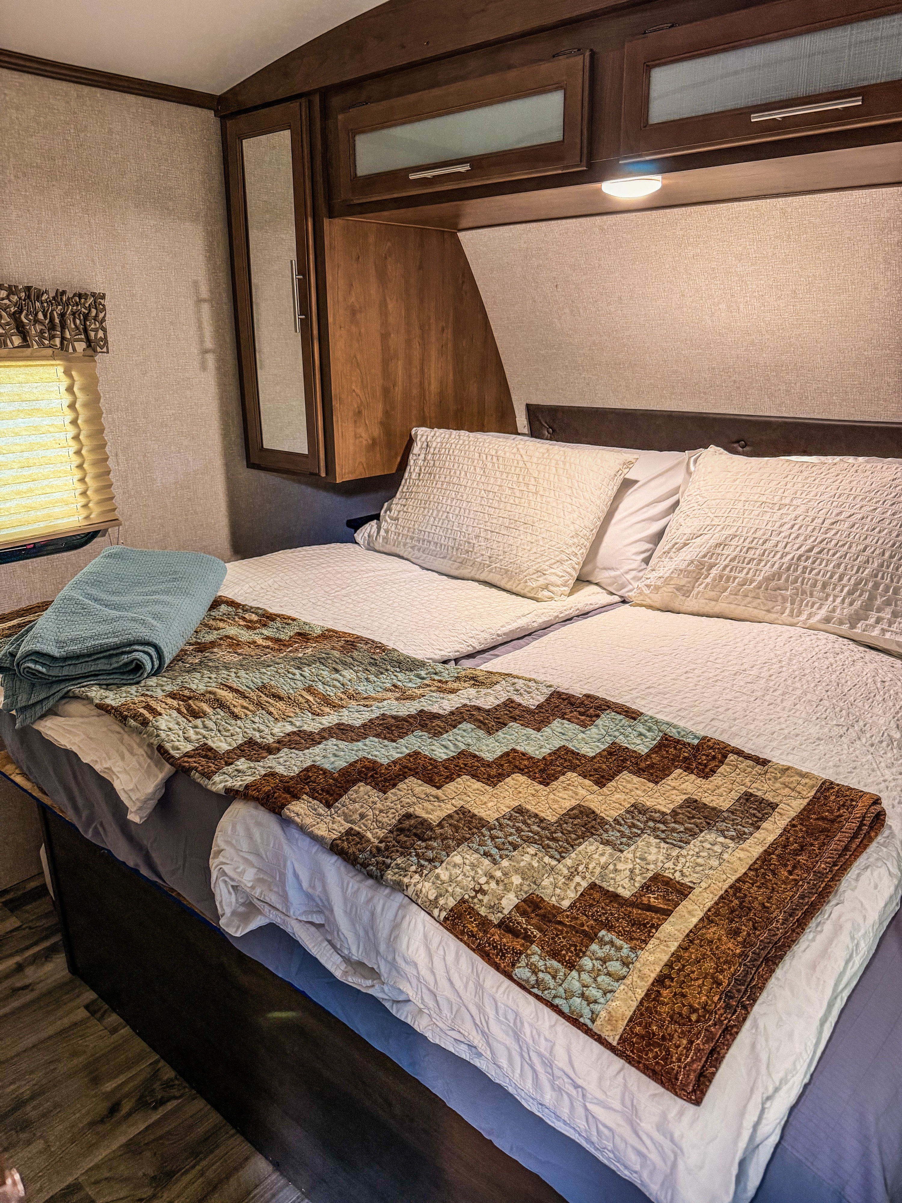 This cozy RV rental bedroom features a neatly made bed with pillows, a chevron quilt, and wood overhead cabinets.. Keystone RV Cougar 2019