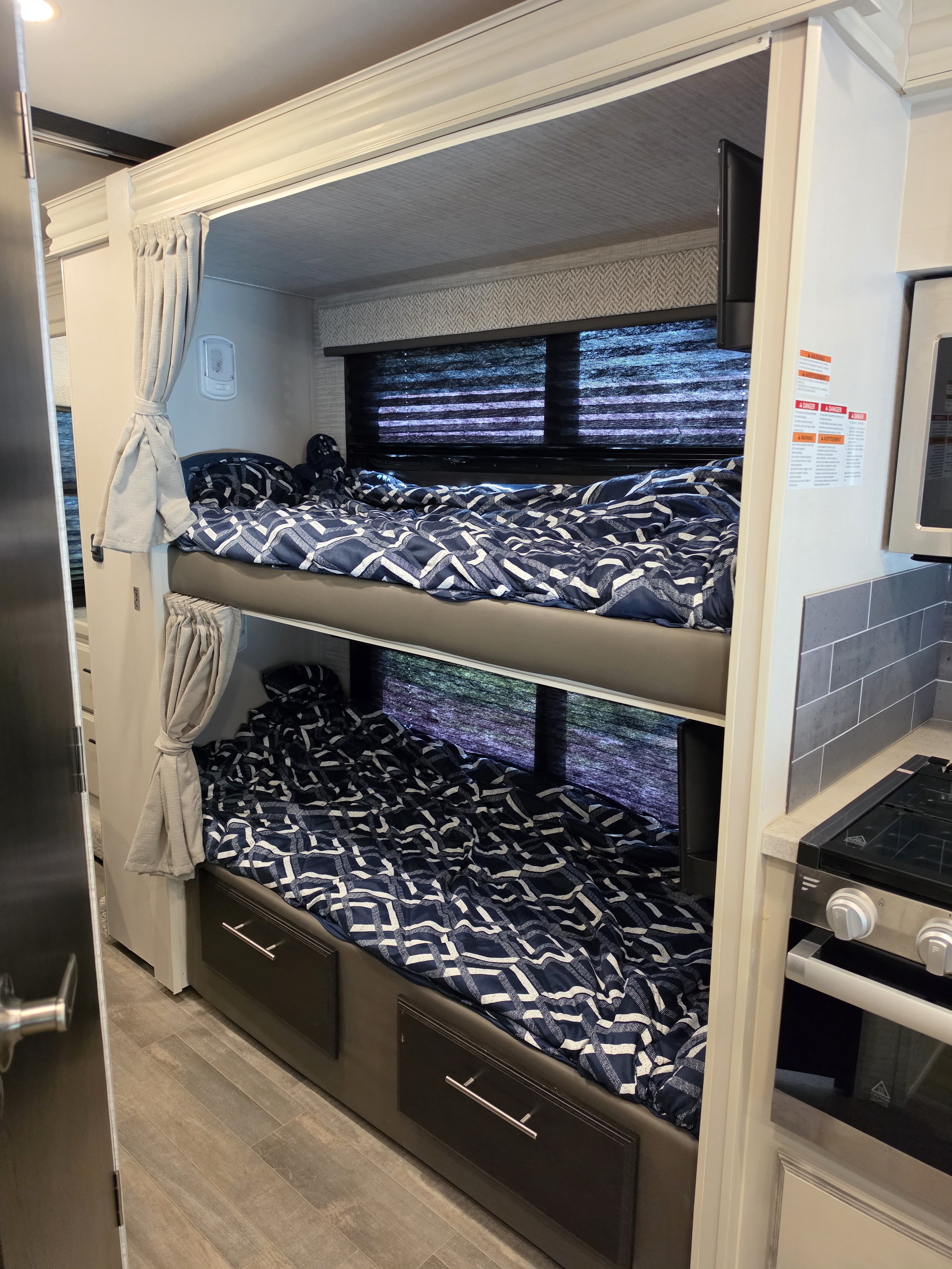 RV rental bunk beds feature navy bedding, tied-back curtains, a window, small TV, and adjacent kitchen area.. Jayco Redhawk 2024