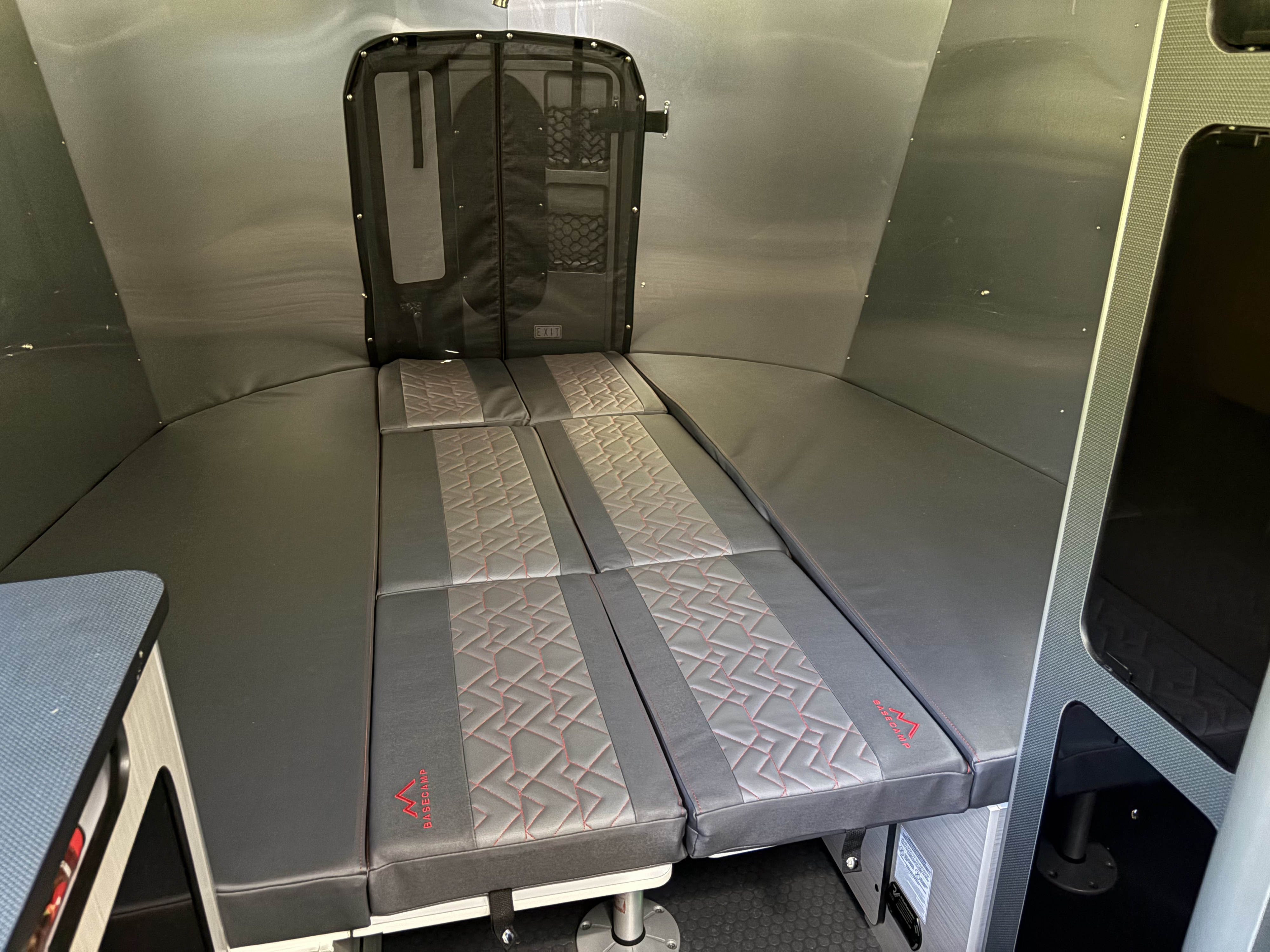 RV rental interior: modular grey & black bed/seating cushions with red geometric pattern and "BASECAMP" logo, metallic walls.. Airstream Basecamp 2025