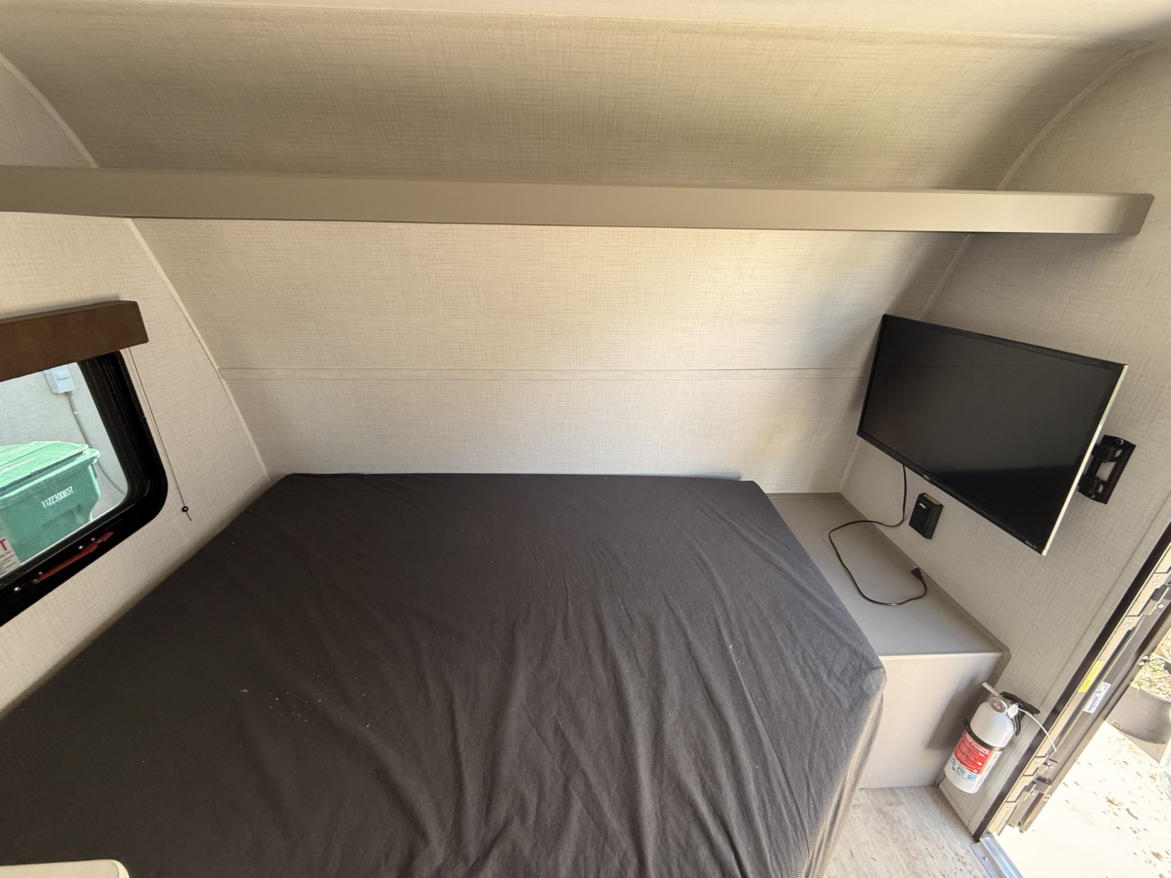 RV rental interior: cozy bedroom with dark bed, mounted TV, window showing outside, overhead shelf, and safety fire extinguisher.. Keystone RV Hideout 2026