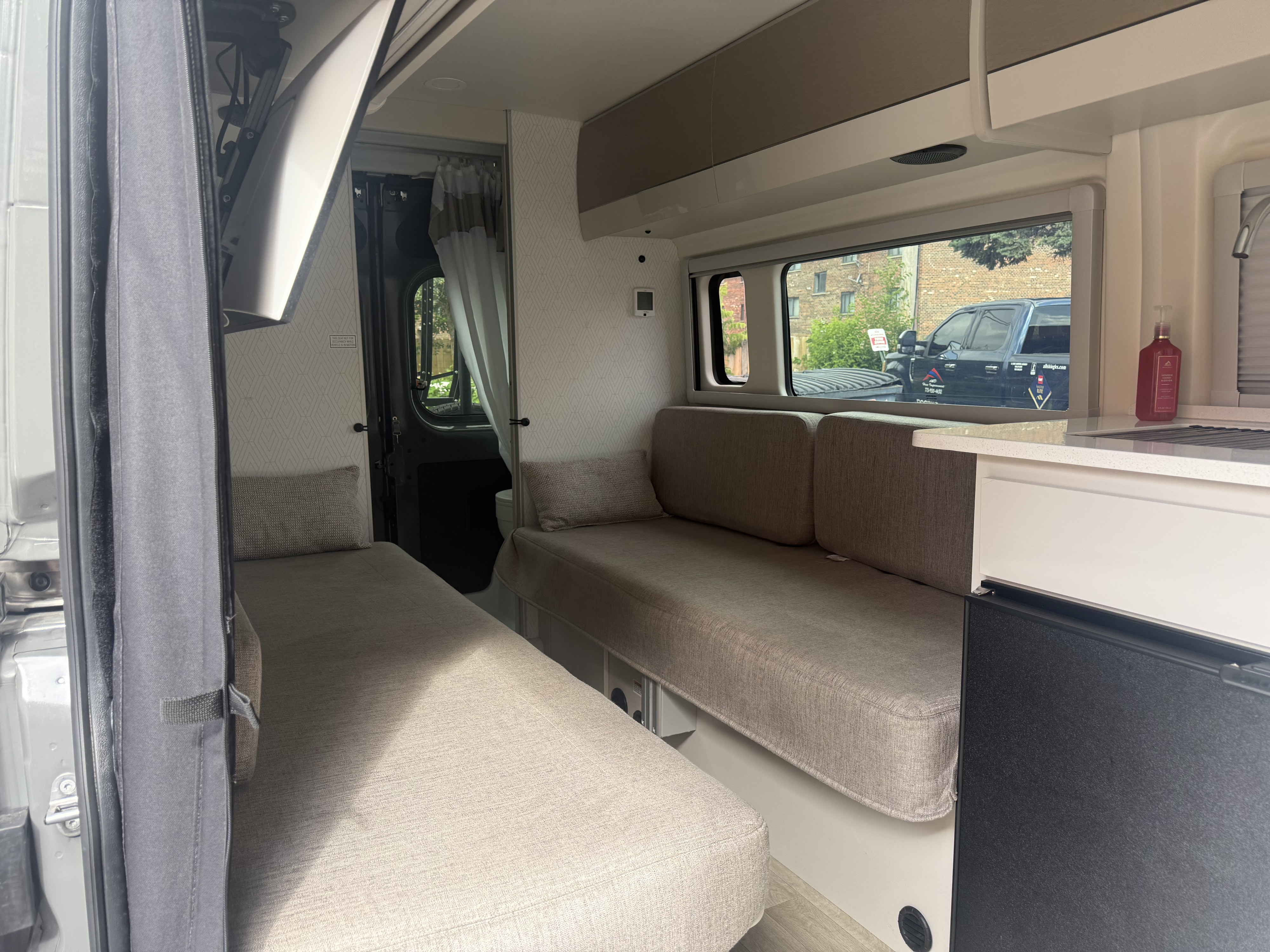RV rental interior: Bright camper van with two convertible sofa beds, a compact kitchenette, and overhead storage.. RAM PROMASTER350 2026