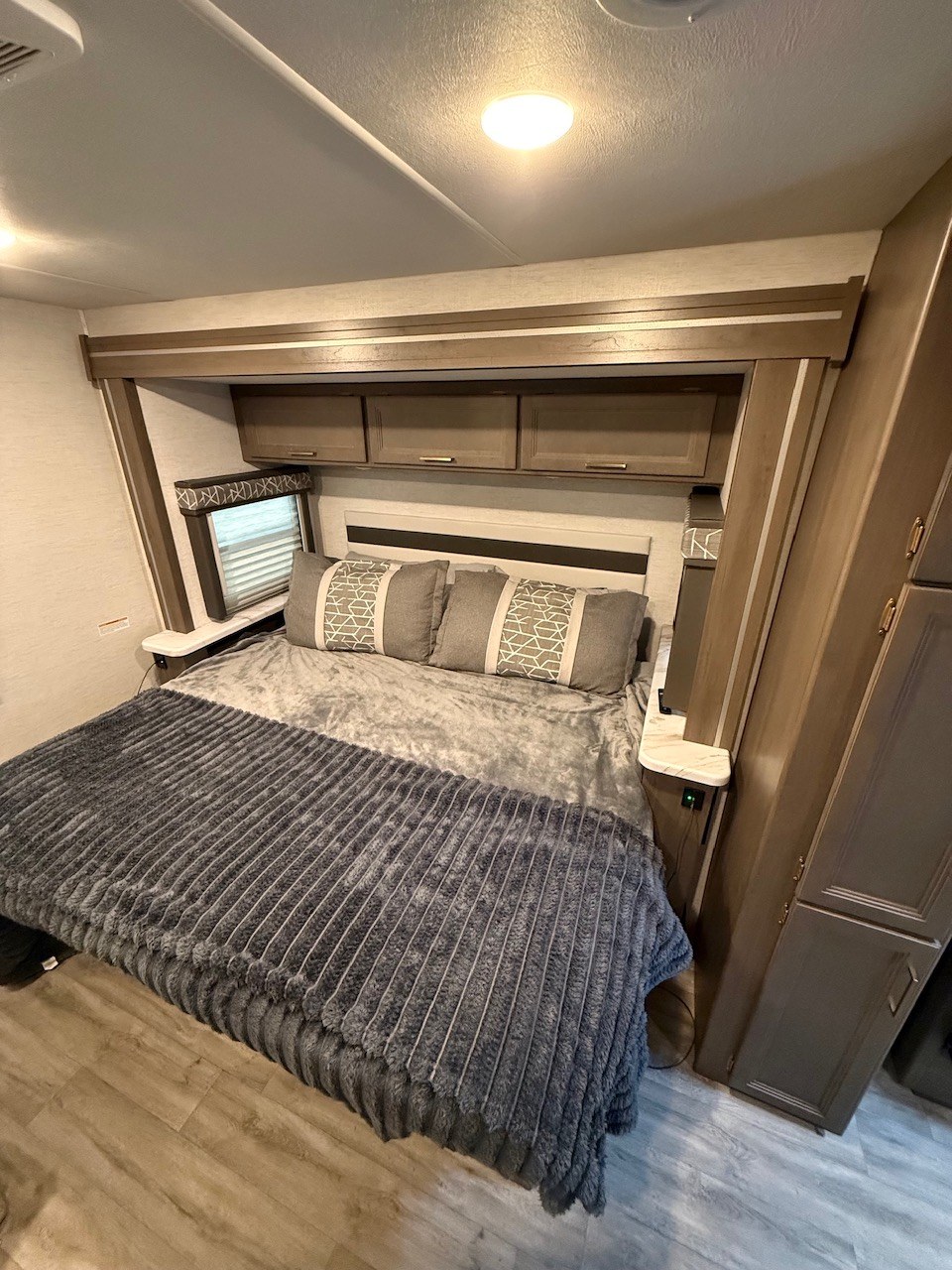 Cozy RV rental bedroom with a large bed, grey bedding, decorative pillows, overhead cabinets, and a small window.. Thor Resonate 2025
