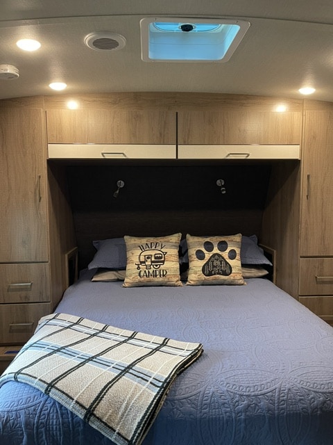 This RV rental features a comfortable bedroom with a blue quilted bed, 'Happy Camper' pillows, overhead storage, and ceiling vent.. Grand Design Imagine 2022