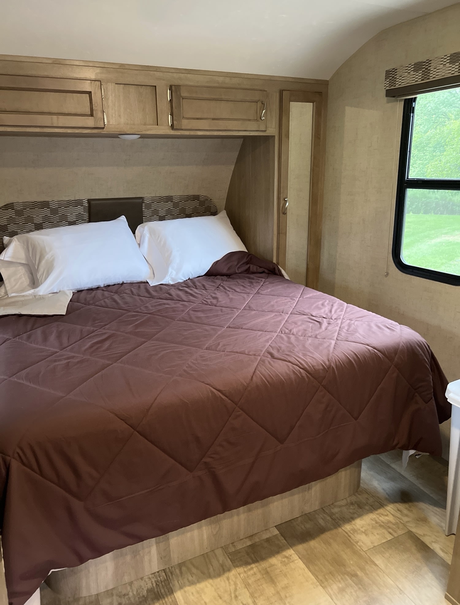 Experience comfort in this RV rental bedroom: brown comforter, white pillows, overhead cabinets, and a window with green views.. KZ VISION 2017