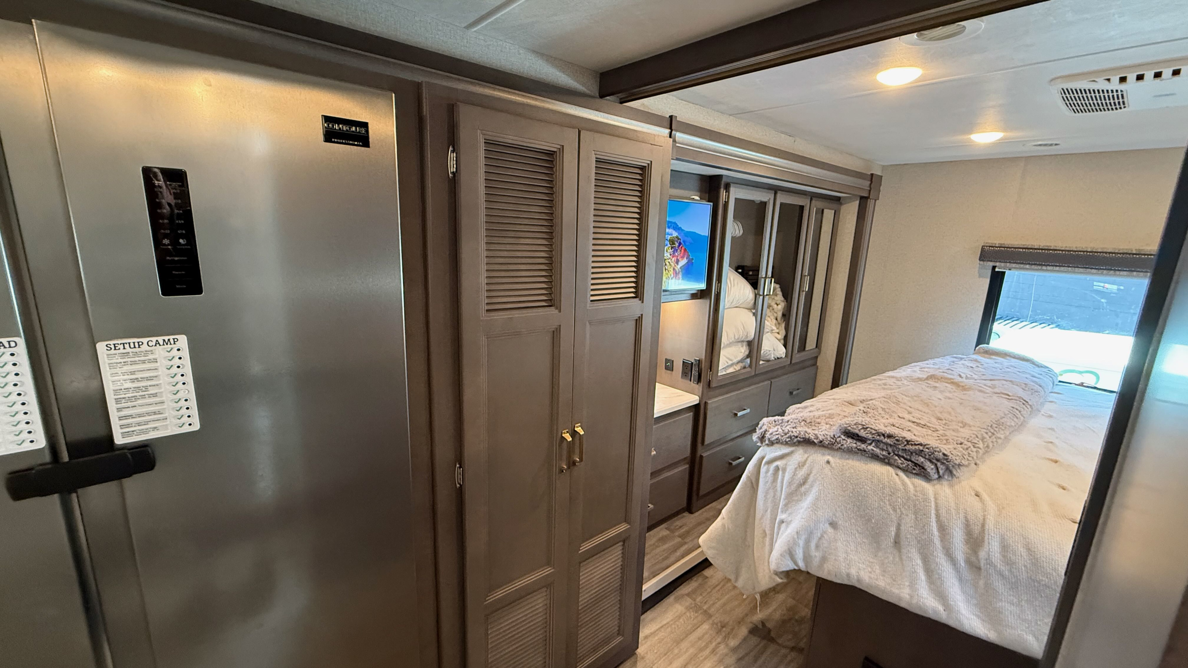 Cozy RV rental interior featuring a modern bedroom with a comfortable bed, large refrigerator, and built-in cabinetry.. Thor Luminate 2024