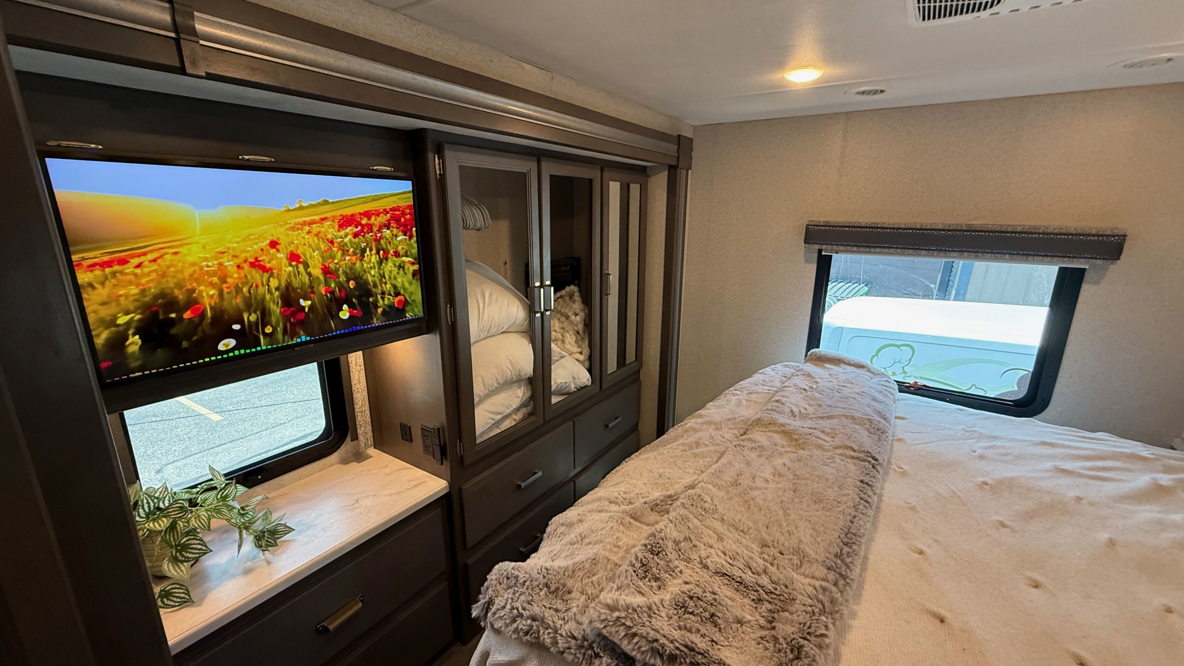 Cozy RV rental bedroom features a TV showing a flower field, plush bedding, and built-in storage with wardrobe and drawers.. Thor Luminate 2024