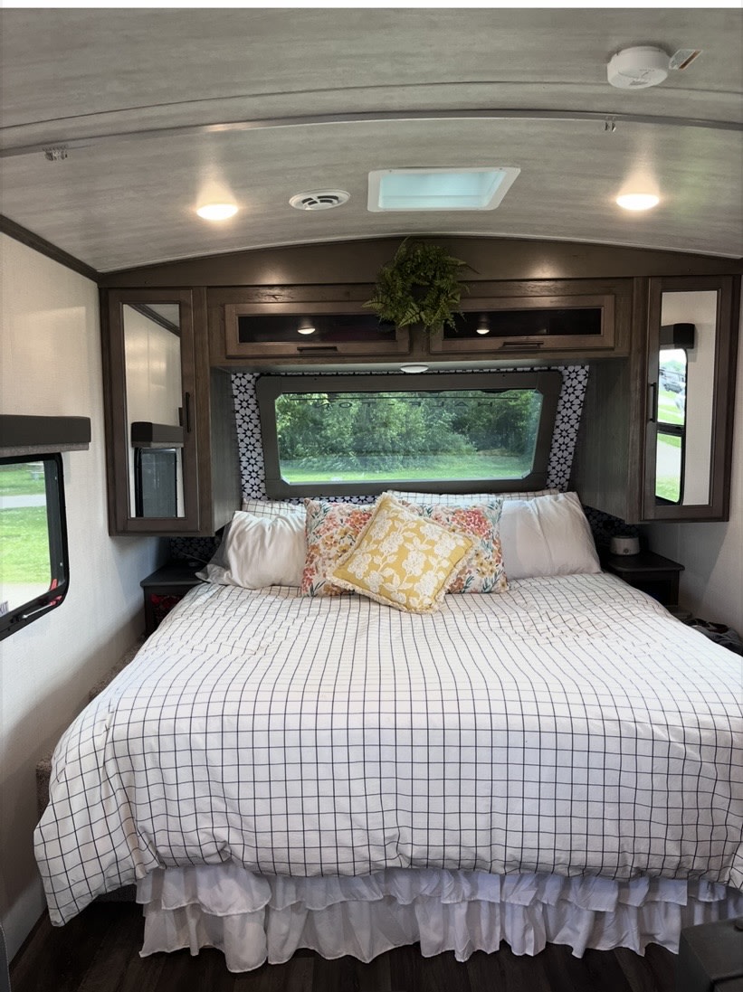 Cozy RV rental bedroom with checkered duvet, floral pillows, overhead storage, and a scenic window view.. Keystone RV Cougar 2023