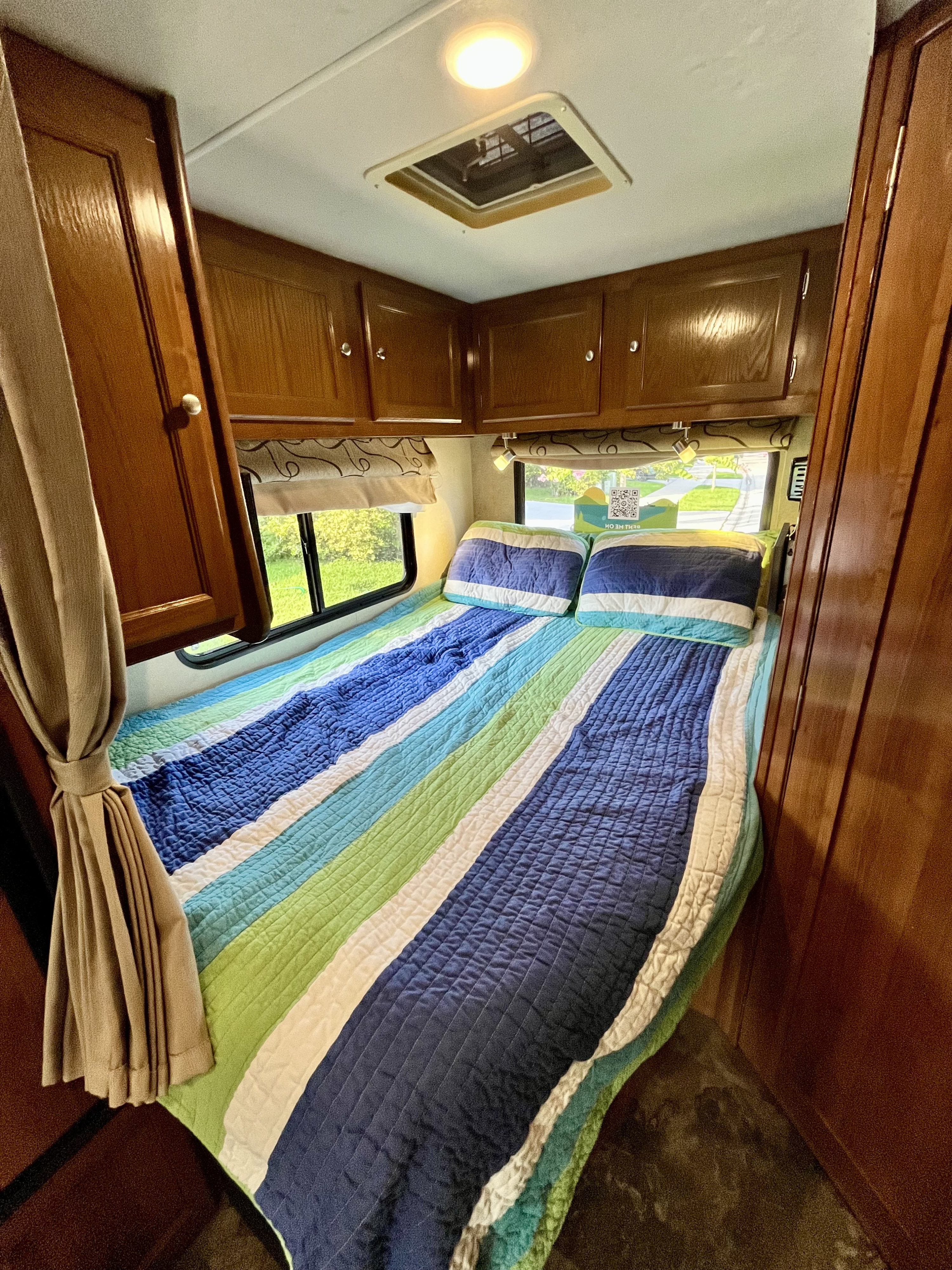 Cozy RV rental bedroom interior featuring a colorful striped bed, windows, and compact overhead storage for a comfortable stay.. Forest River Coachmen Leprechaun 2019