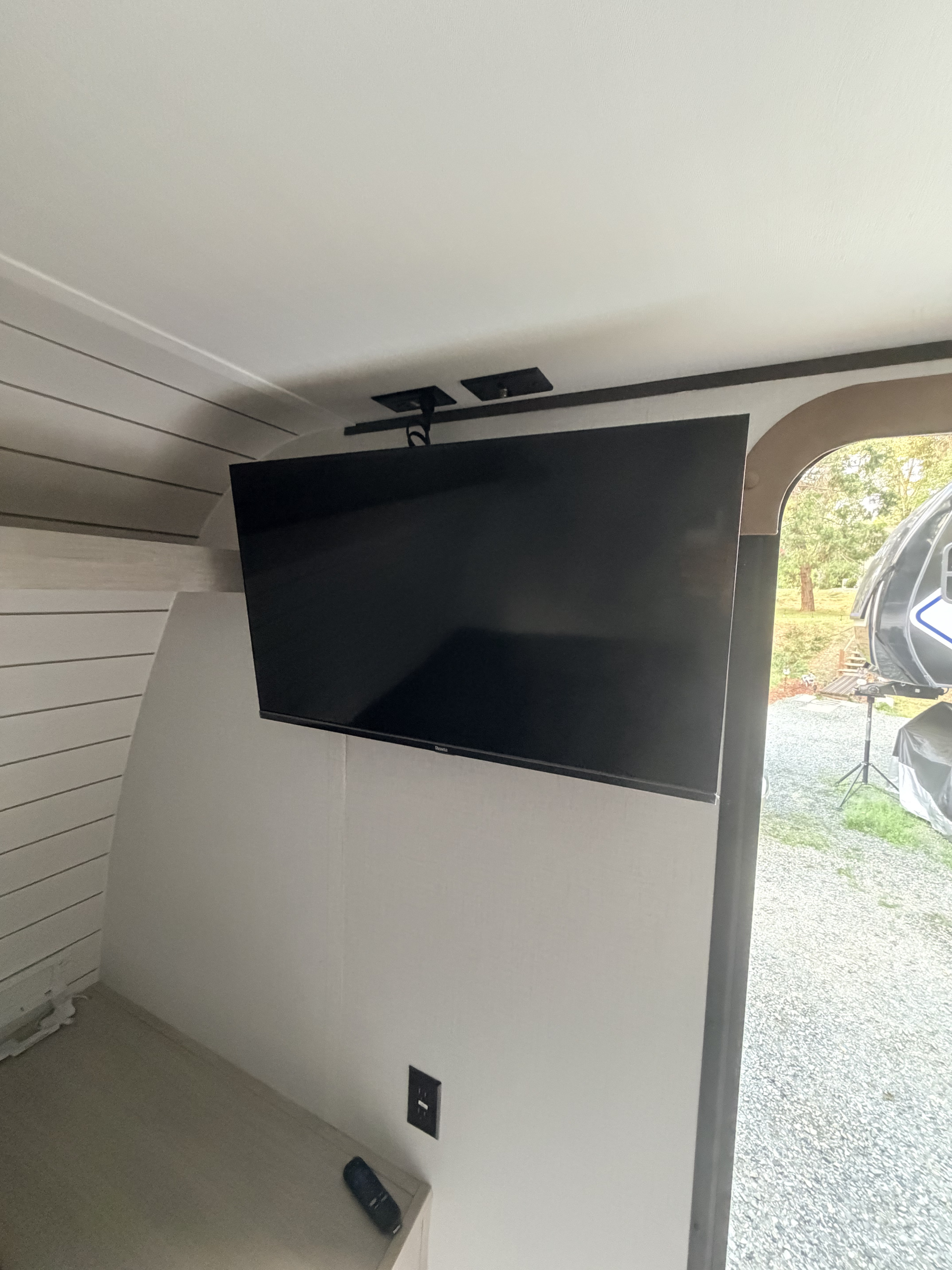 RV rental interior: A ceiling-mounted Roku TV hangs over a counter with a remote and outlet. Gravel yard visible outside.. Keystone RV Crossrads Zinger 18BH 2026