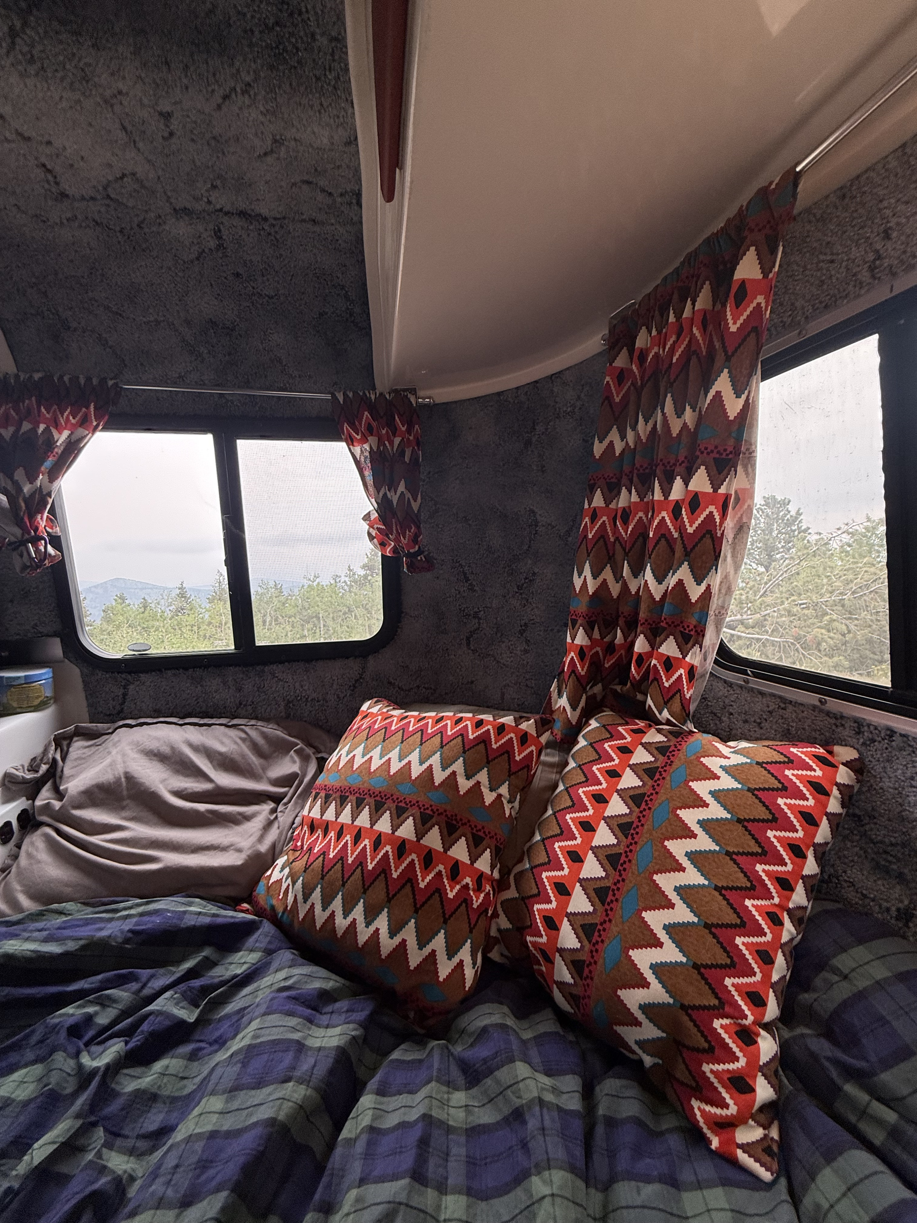 Cozy RV rental interior with a bed, colorful patterned pillows & curtains, and windows looking out to a mountain forest view.. Casita 16 1986