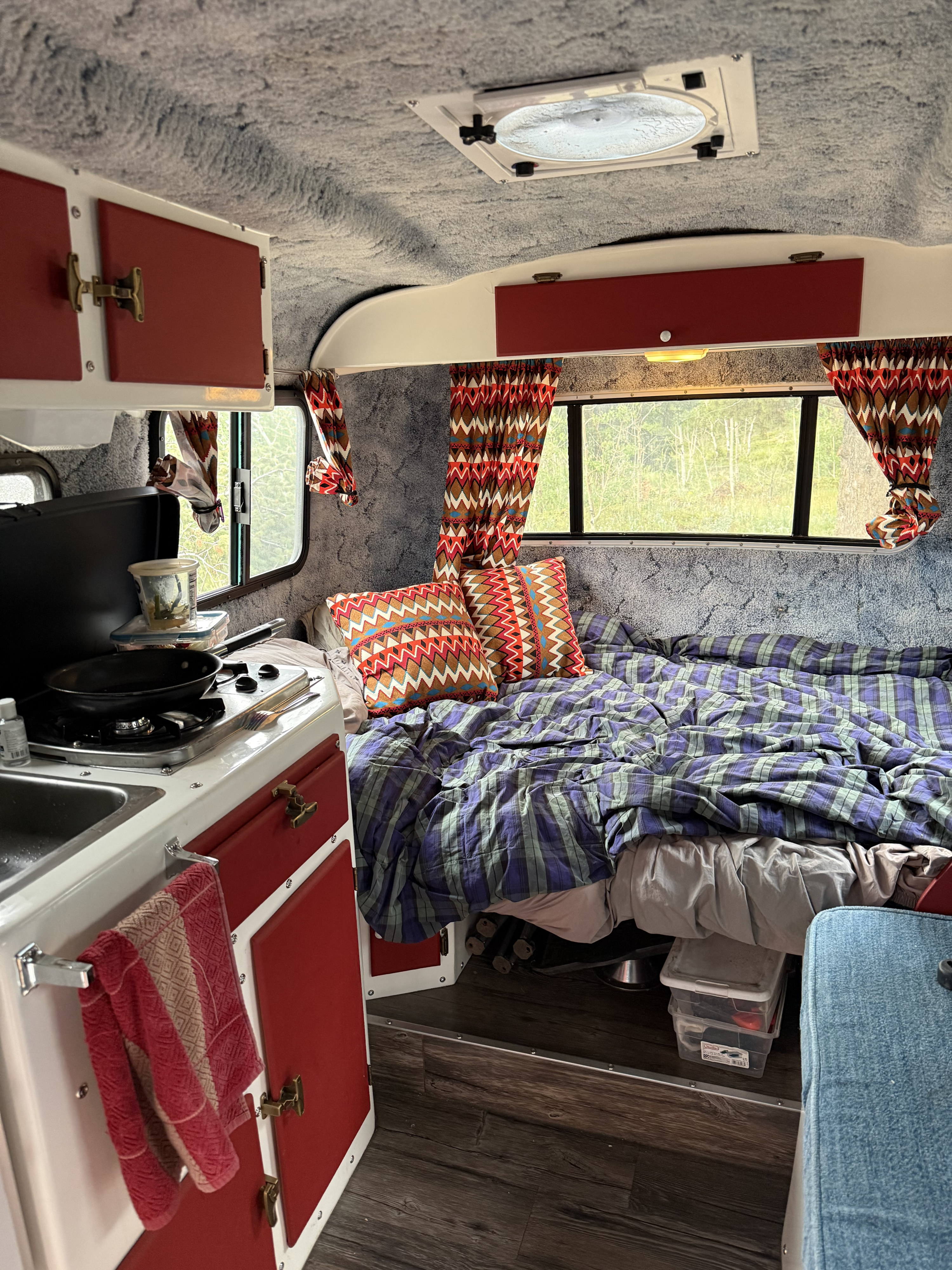 An inviting RV rental interior, showcasing a comfy bed with plaid comforter, compact kitchenette, and window view.. Casita 16 1986