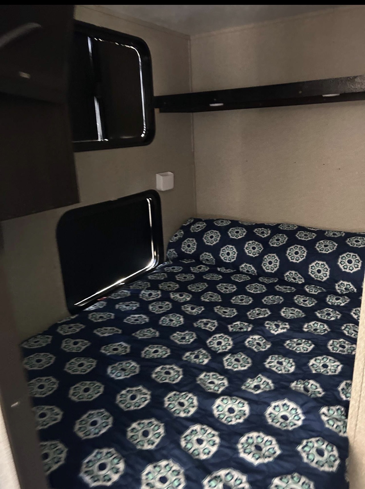 Cozy sleeping area in an RV rental, featuring a bed with a blue patterned comforter, windows, and a shelf.. Keystone RV Springdale Eco-Lite 2019