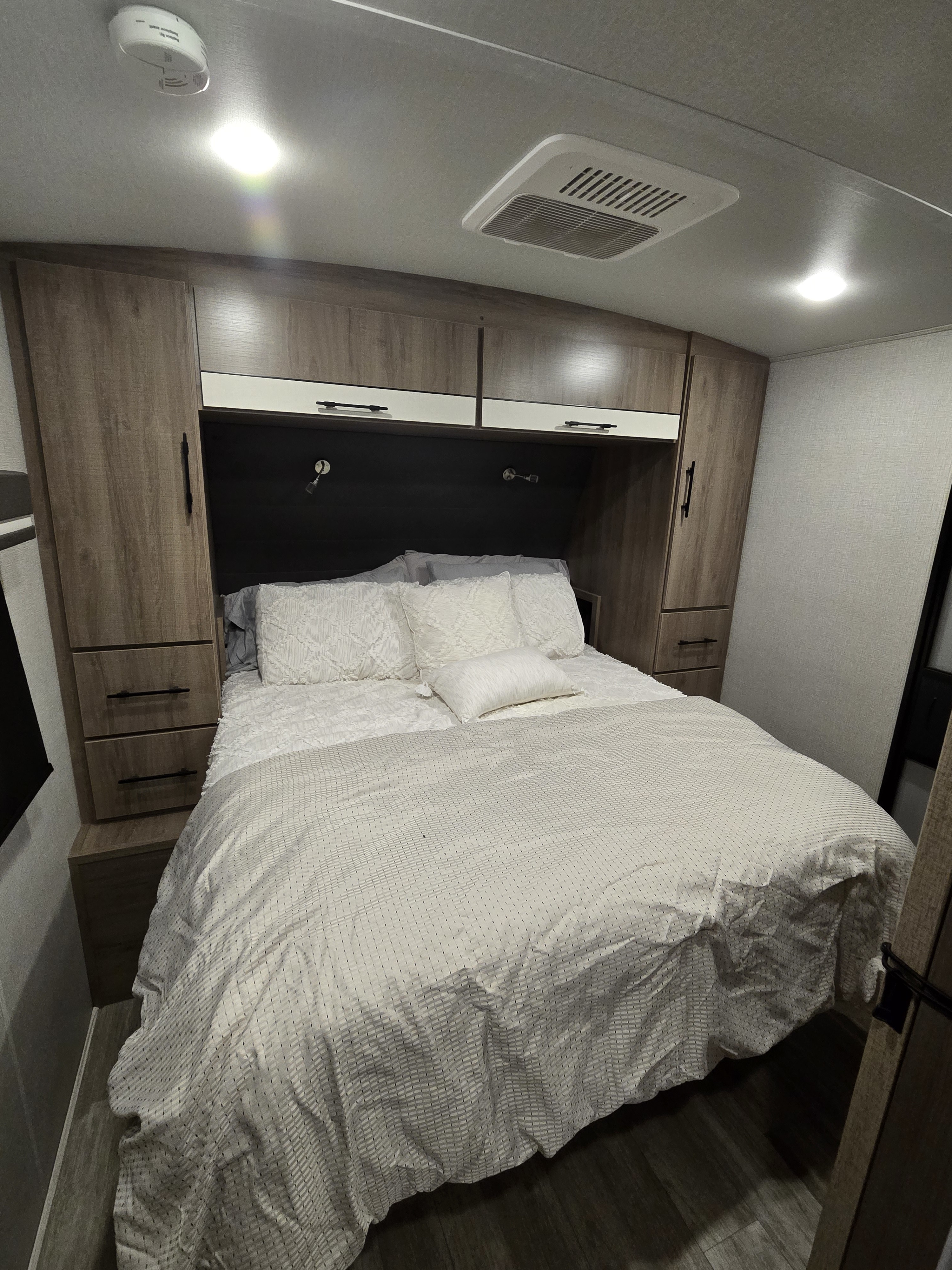 Cozy RV rental bedroom featuring a comfortable bed with white bedding, wooden cabinets, and overhead lighting with an AC vent.. Grand Design Imagine 2023