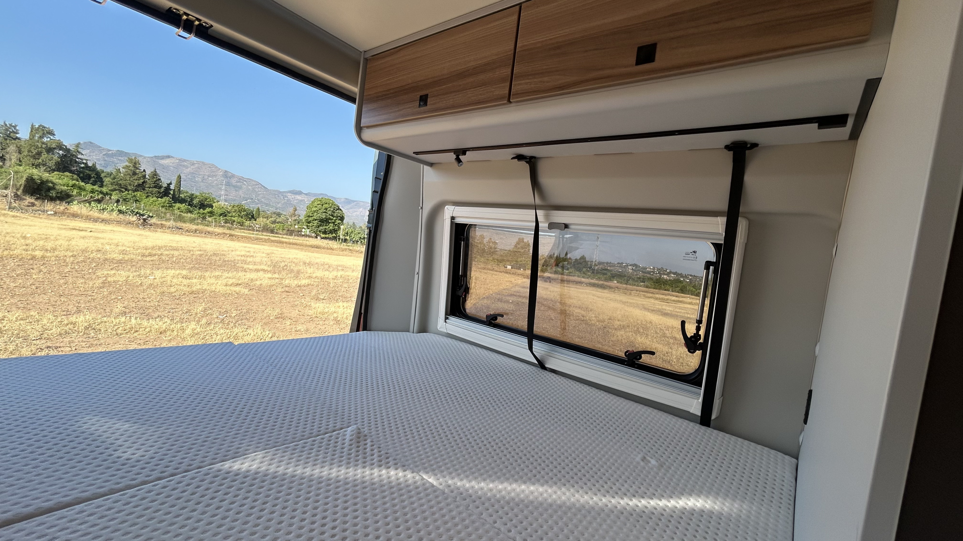 Inside an **RV rental**: a comfortable bed facing a window with a beautiful natural view of dry fields and distant mountains.. BURSTNER B66 2024