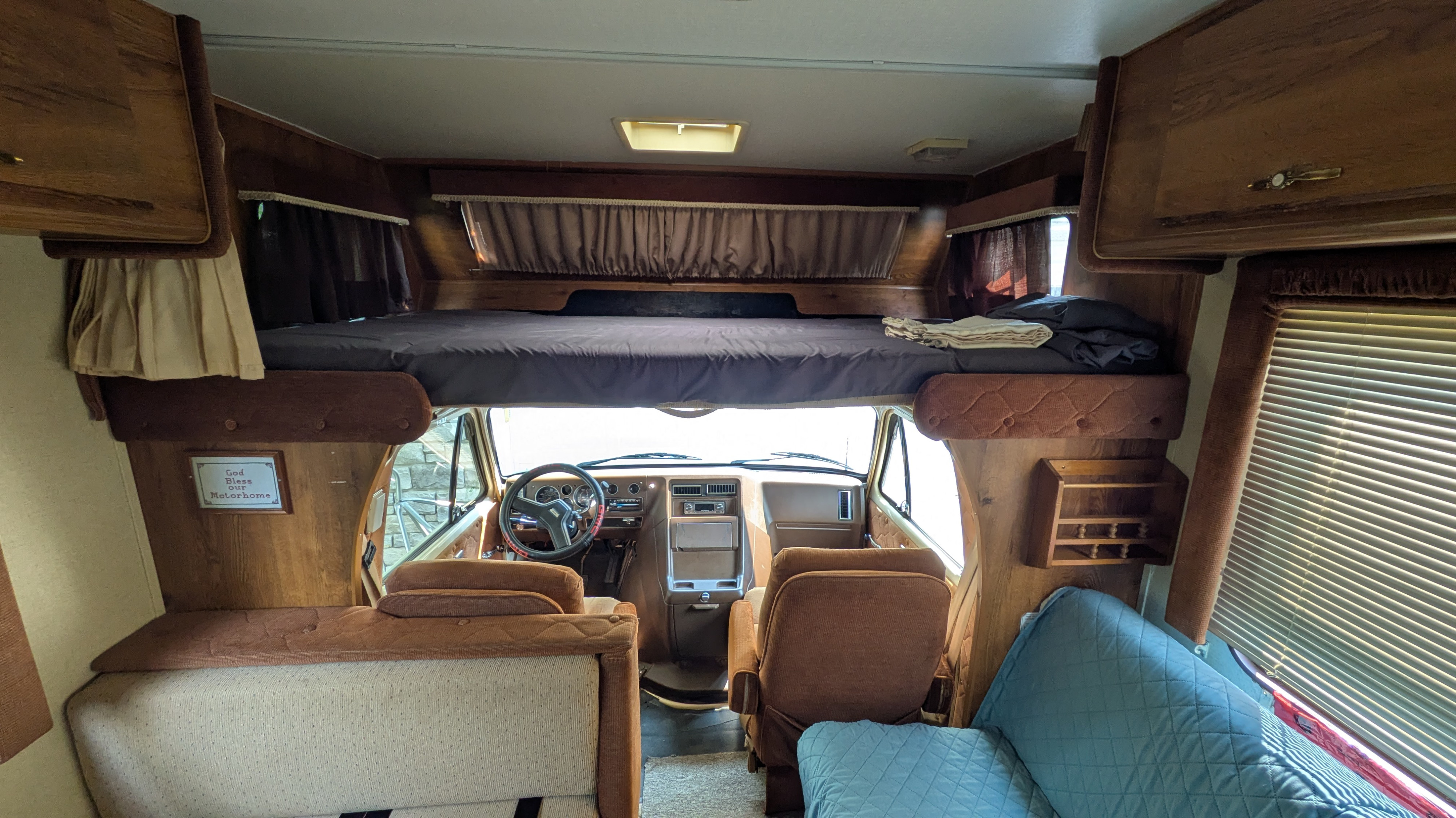 Inside a vintage rv rental, showing the driver's area, an overhead bed, and dinette. Warm wood tones throughout.. Gmc Motorhome 1987