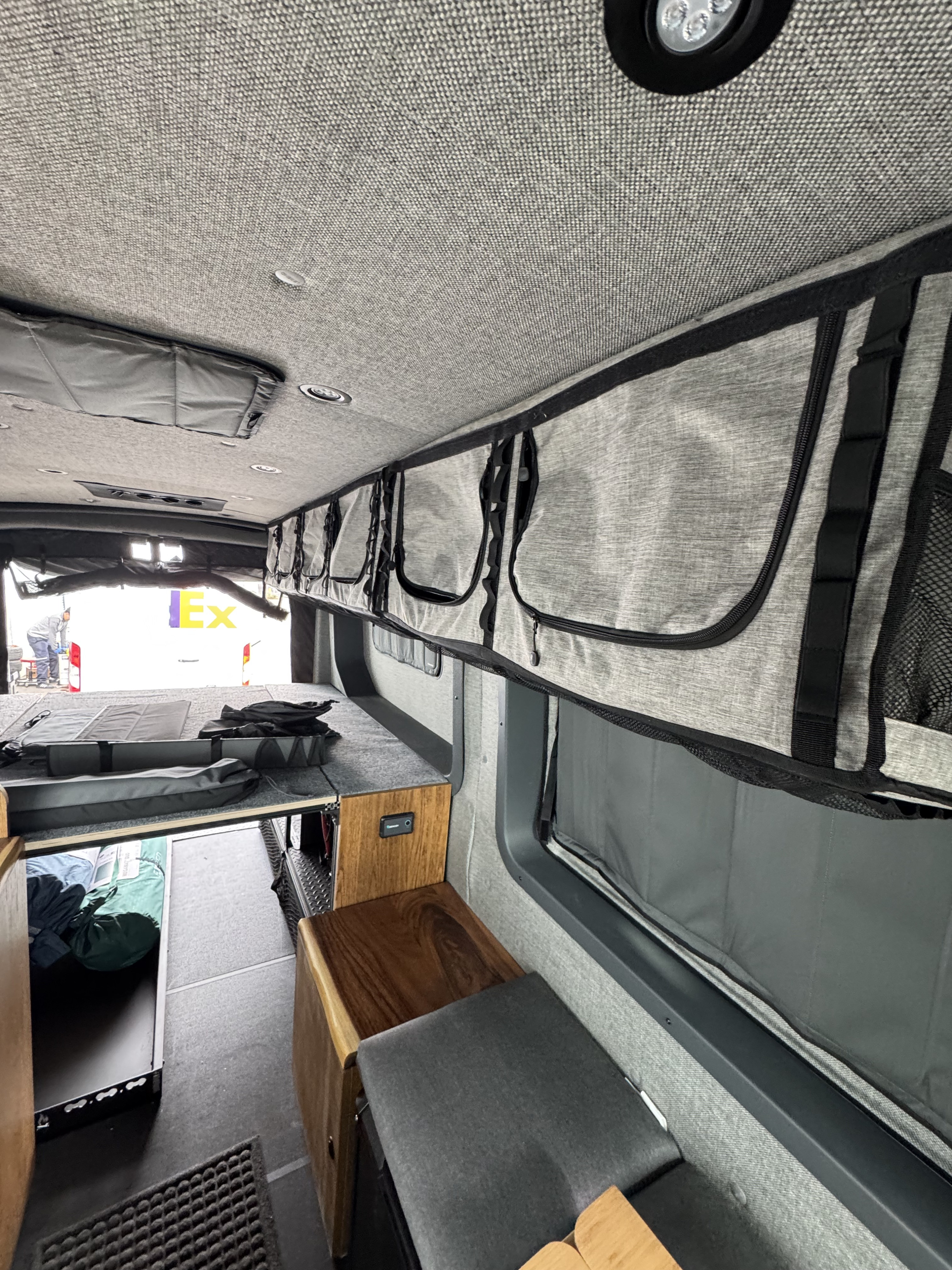 RV rental camper van interior. Grey fabric walls/ceiling, modular overhead storage, bed platform, and wood accents.. Ford Transit 2024