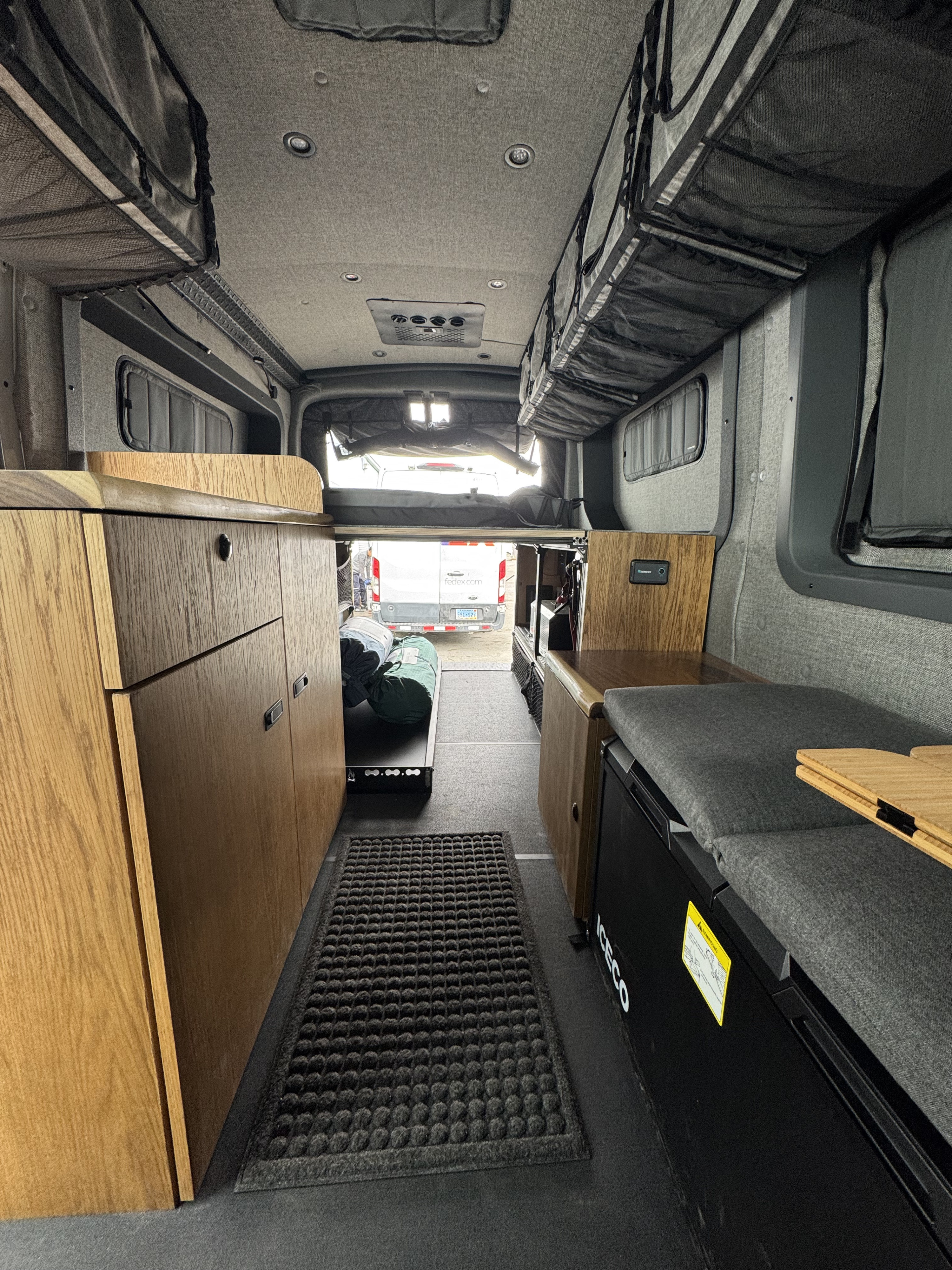 Interior of an RV rental camper van with light wood cabinets, an Iceco fridge, bed, and mesh ceiling storage. A cozy mobile home.. Ford Transit 2024